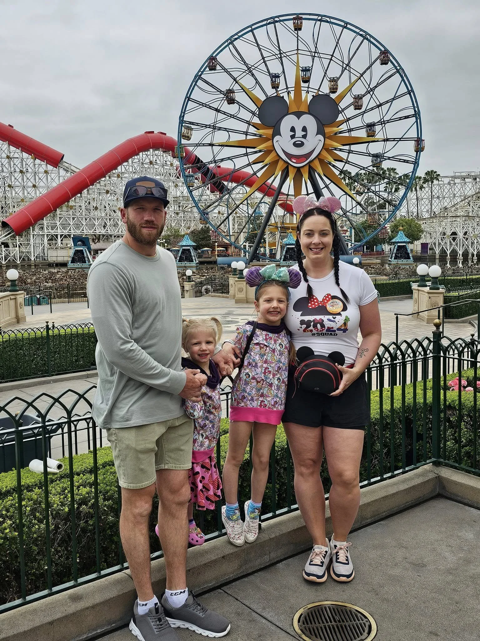 A family of four standing in front of a Disney California Adventure amusement park ride with a large Mickey Mouse face and a Ferris wheel behind them. The family includes a man, a woman, and two young girls, all smiling. The family is wearing casual clothing and Disney-themed accessories, with the woman wearing Mickey Mouse ears and the younger girl wearing Minnie Mouse ears. The background includes the park's rides and a cloudy sky.