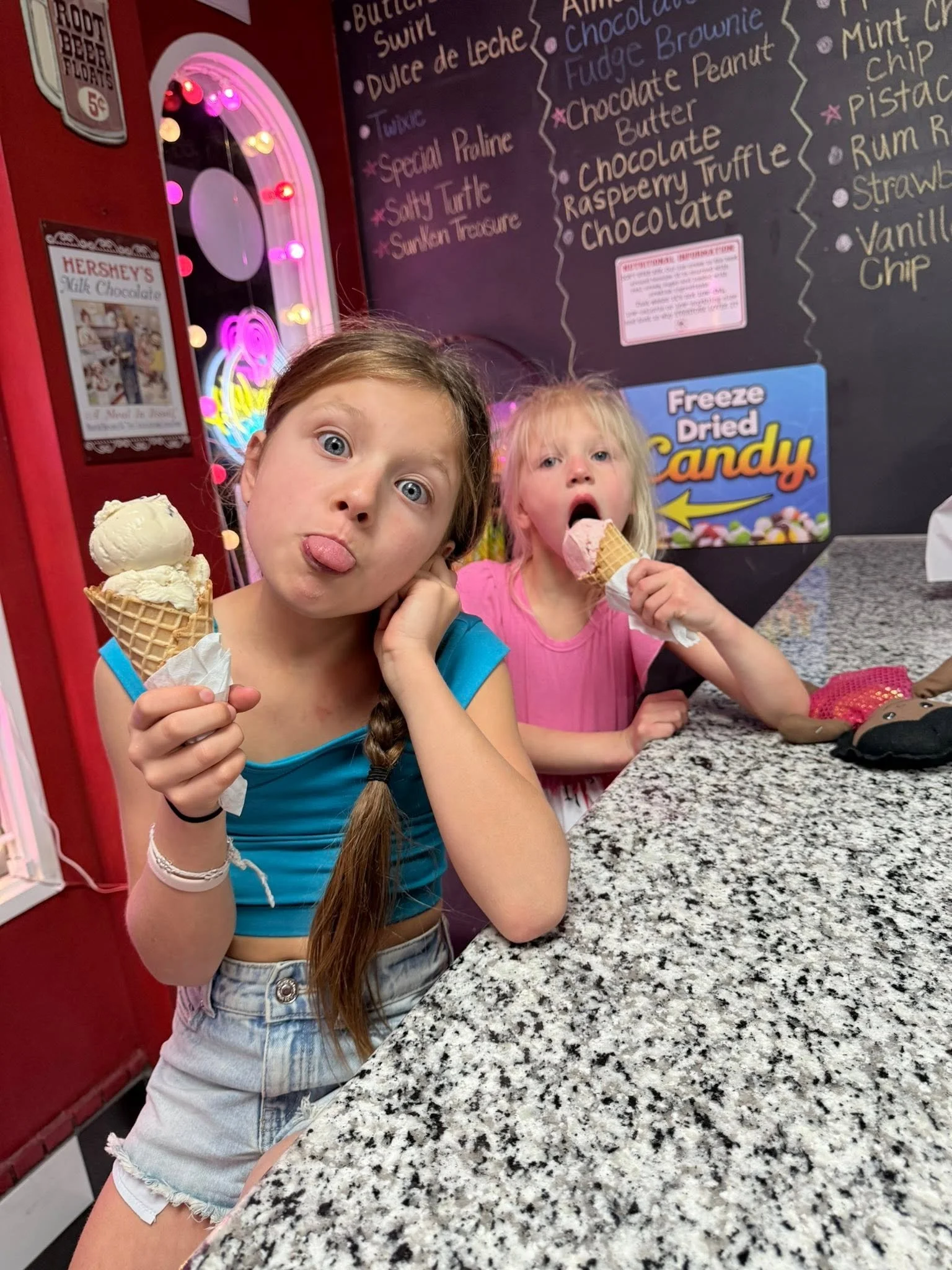 Two young girls with ice cream cones in a dessert shop, one making a silly face and the other eating ice cream, with a menu board in the background and a granite countertop in the foreground.
