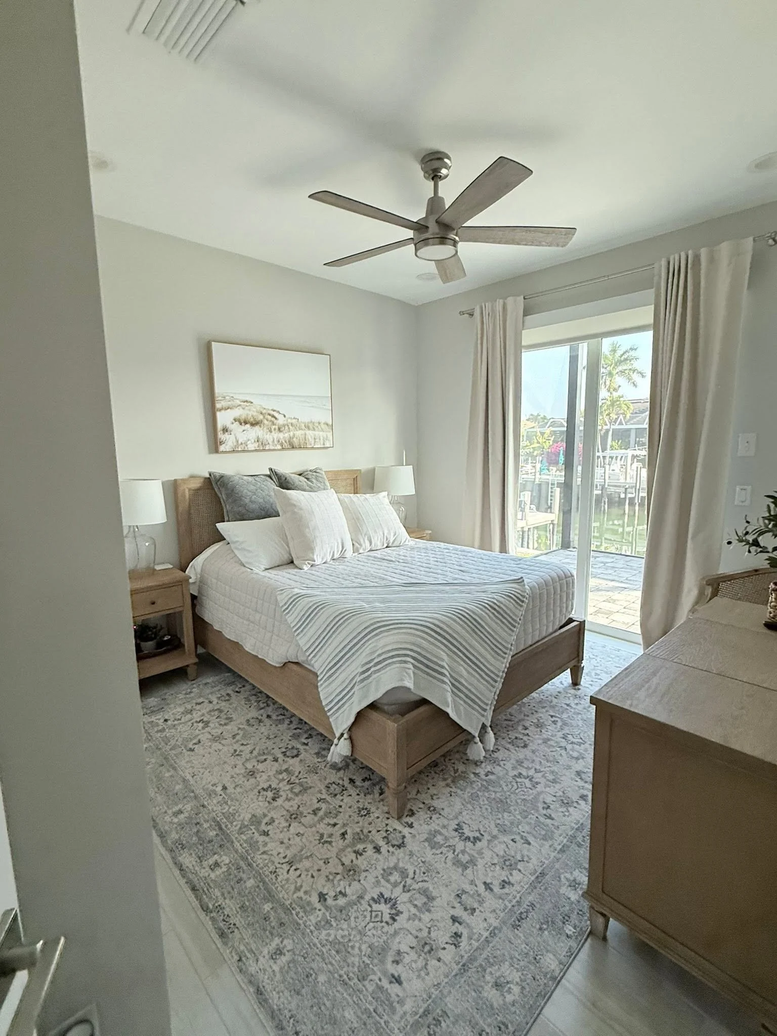 A bedroom with a wooden bed frame, white bedding, and multiple pillows. There are two nightstands with lamps, a decorative rug, and a dresser. A sliding glass door with curtains leads outside, revealing a patio and palm trees.