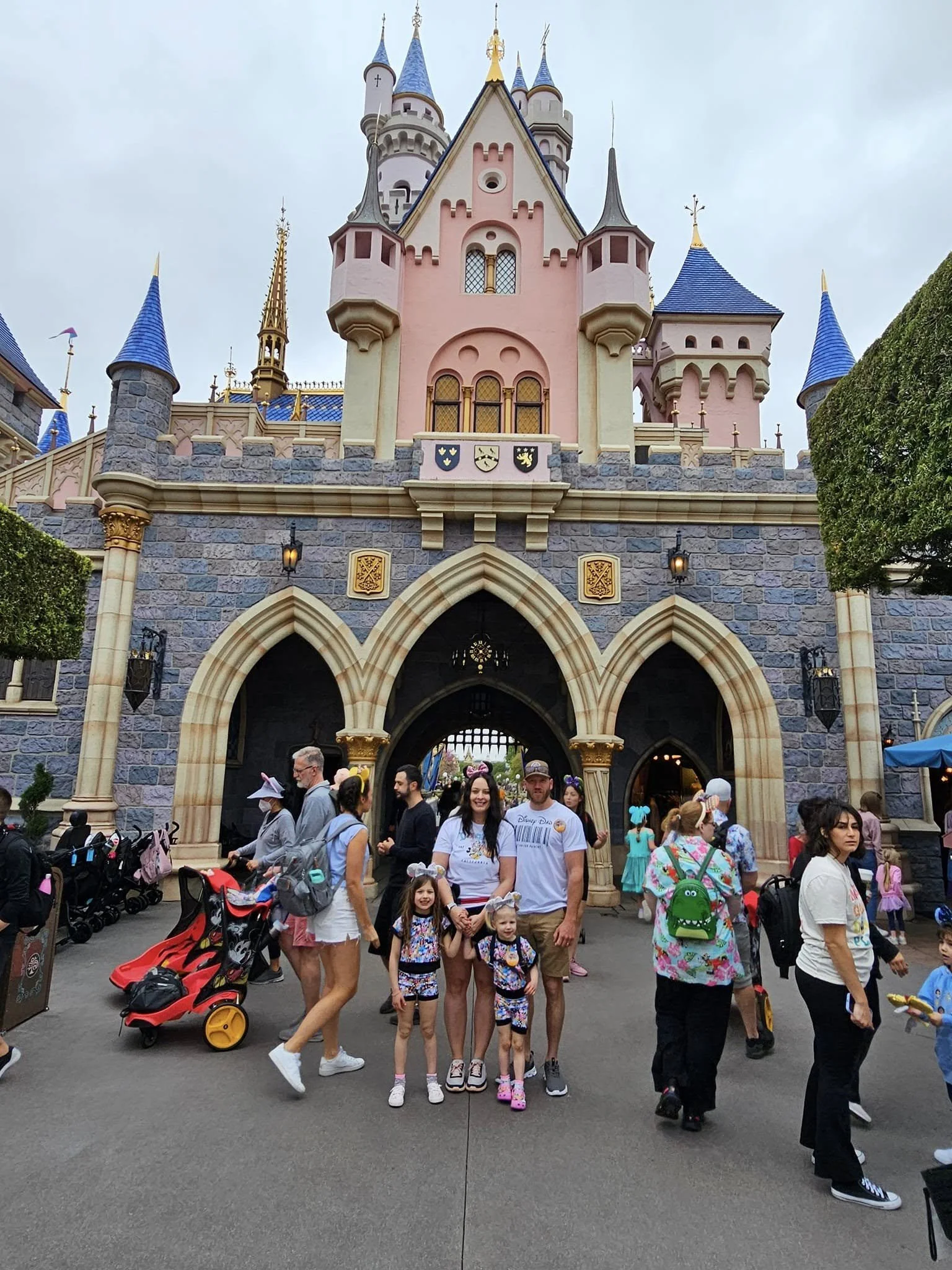 Disneyland castle with visitors in front, some wearing Disney themed costumes and accessories, overcast sky