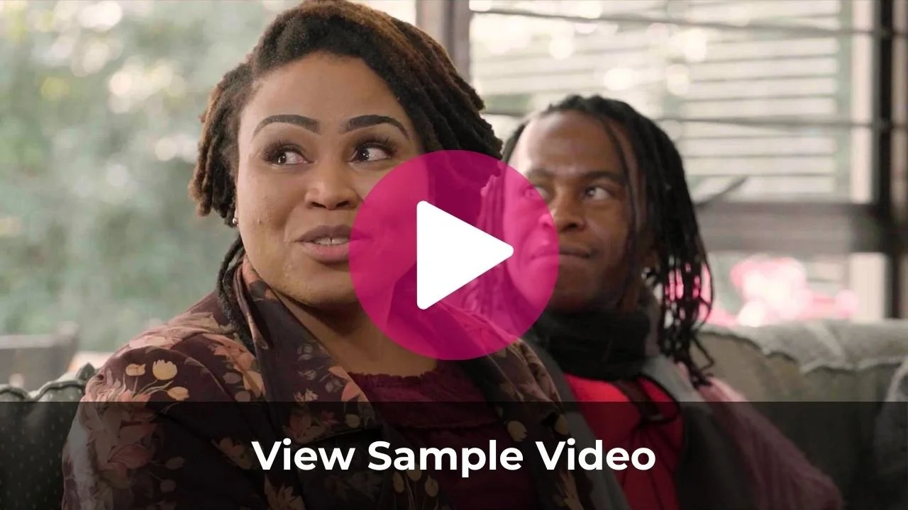 A woman with dreadlocks and makeup sitting on a couch, talking, with a man with dreadlocks in the background looking at her, in a brightly lit room.