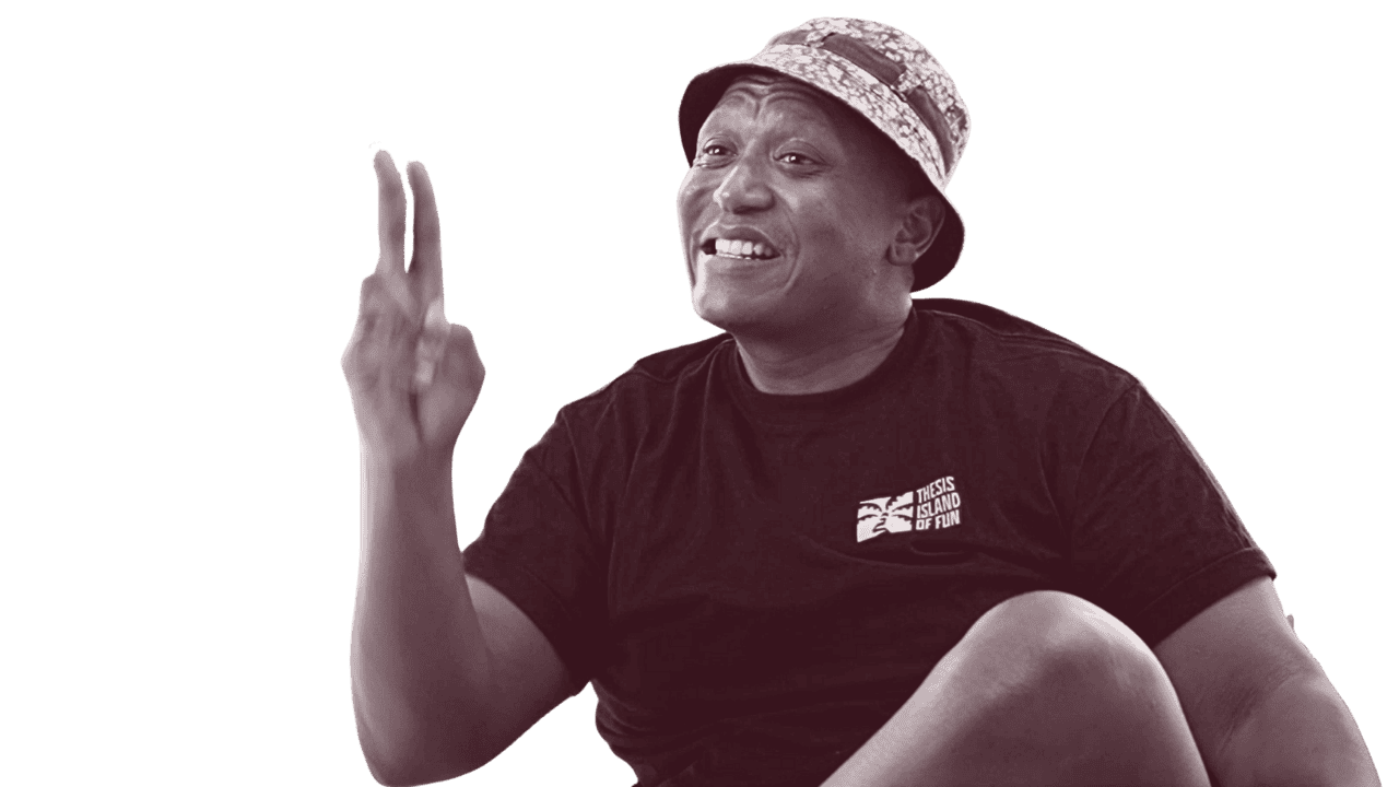 Smiling man wearing a bucket hat and a black T-shirt with a logo, making a peace sign with his right hand against a black background.