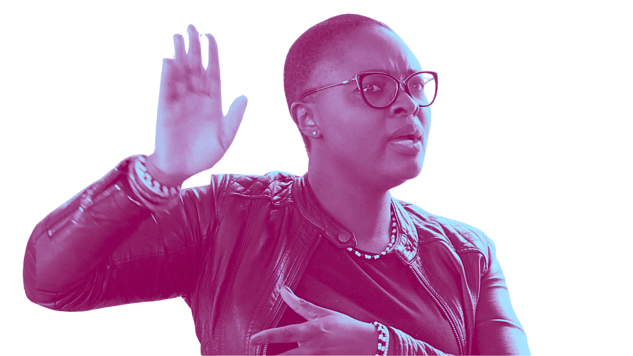 Woman wearing glasses and a leather jacket, raising her right hand and pointing with her left index finger, against a black background.