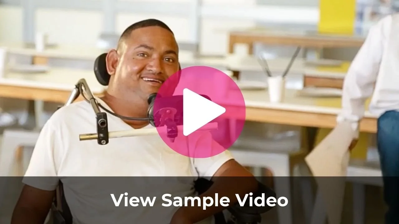 A man using a motorized wheelchair with head and neck support, smiling, in a bright, modern workspace with tables and supplies in the background.