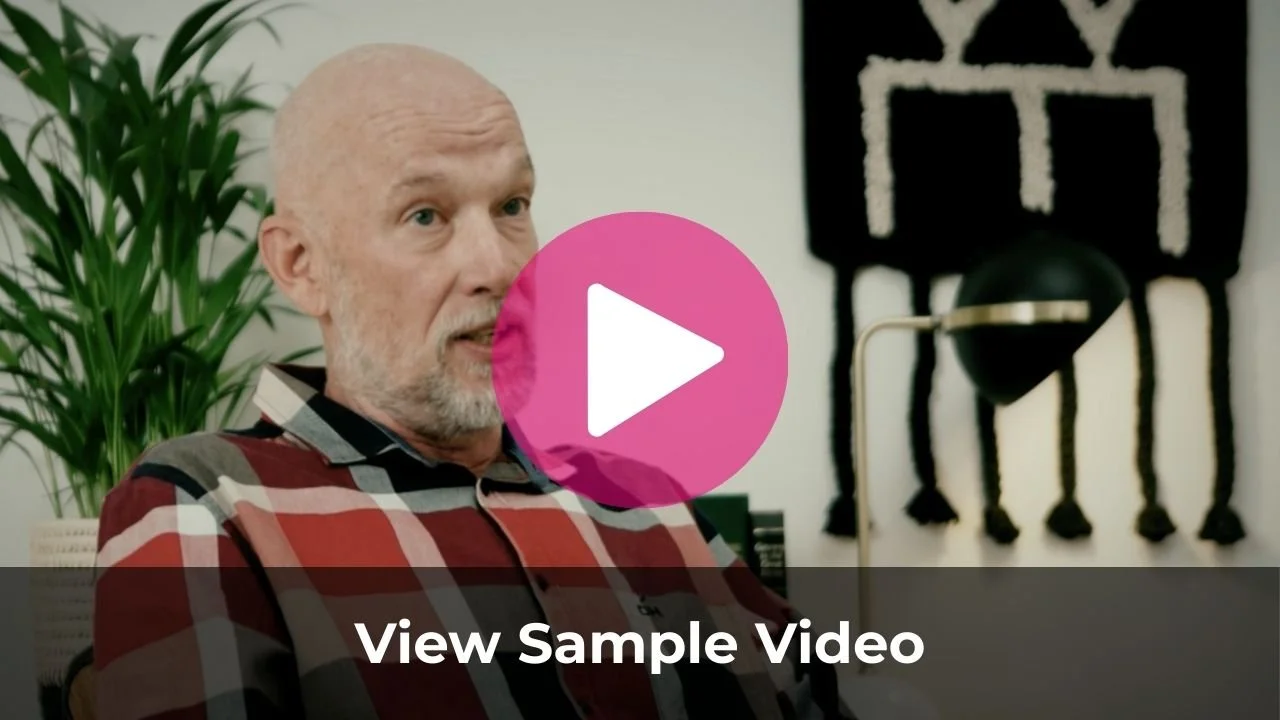 A bald man with a gray beard wearing a red and black plaid shirt sitting in an office, with a green plant and abstract black and white wall art in the background.