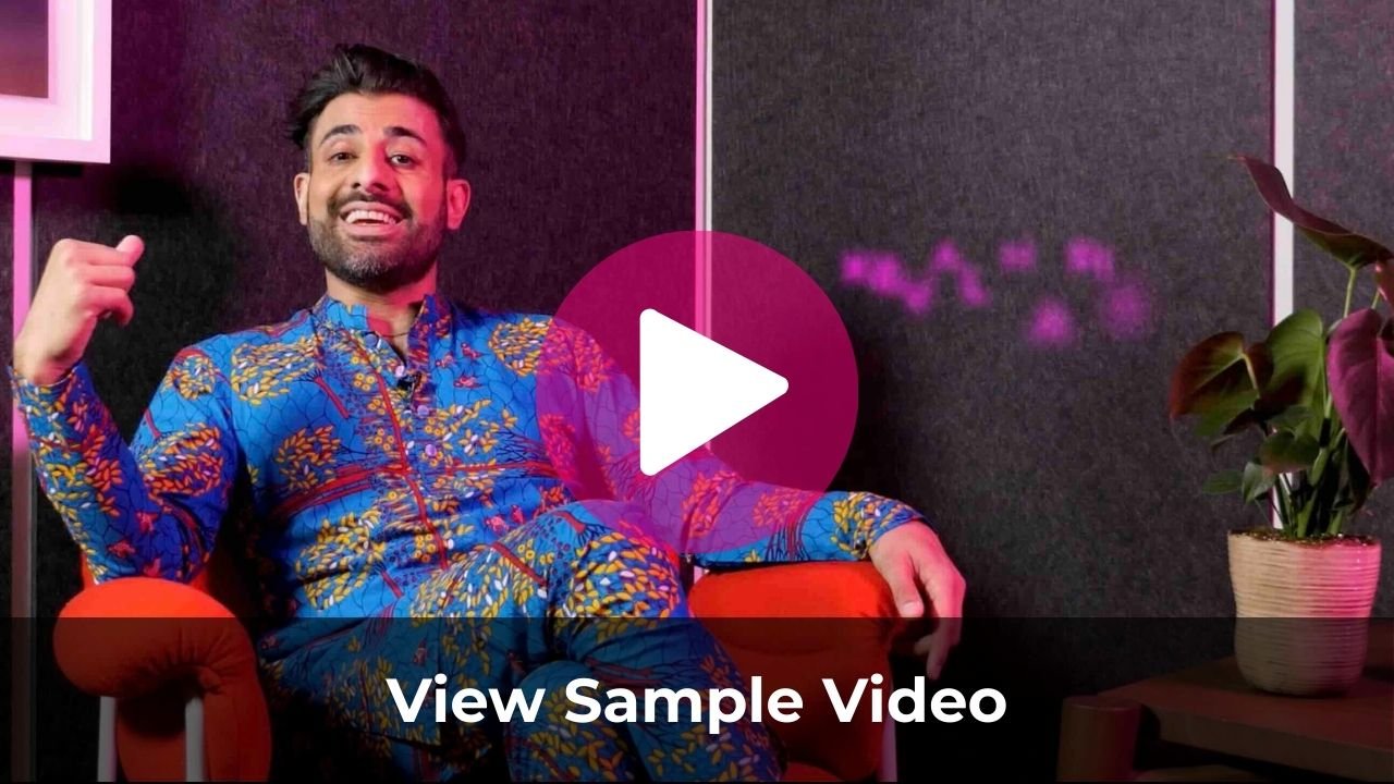A man with dark hair and a beard, wearing a colorful blue and yellow patterned shirt, sitting on a red chair, smiling, with a dark wall and a potted plant nearby.