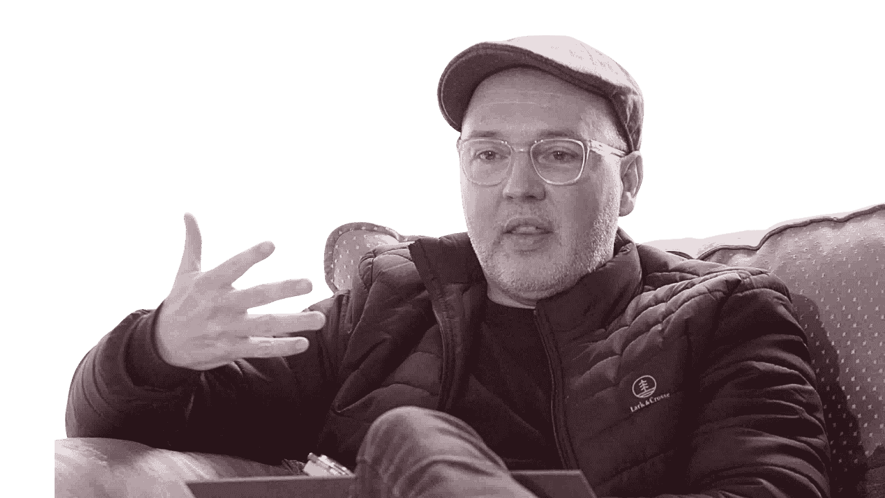 A man with glasses, a cap, and a jacket sitting on a sofa, gesturing with his right hand.