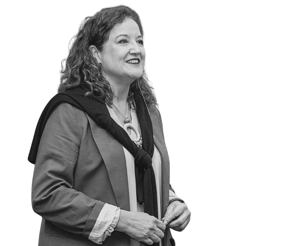 A smiling woman with curly hair, wearing a blazer, a necklace, and a sweater draped around her shoulders.