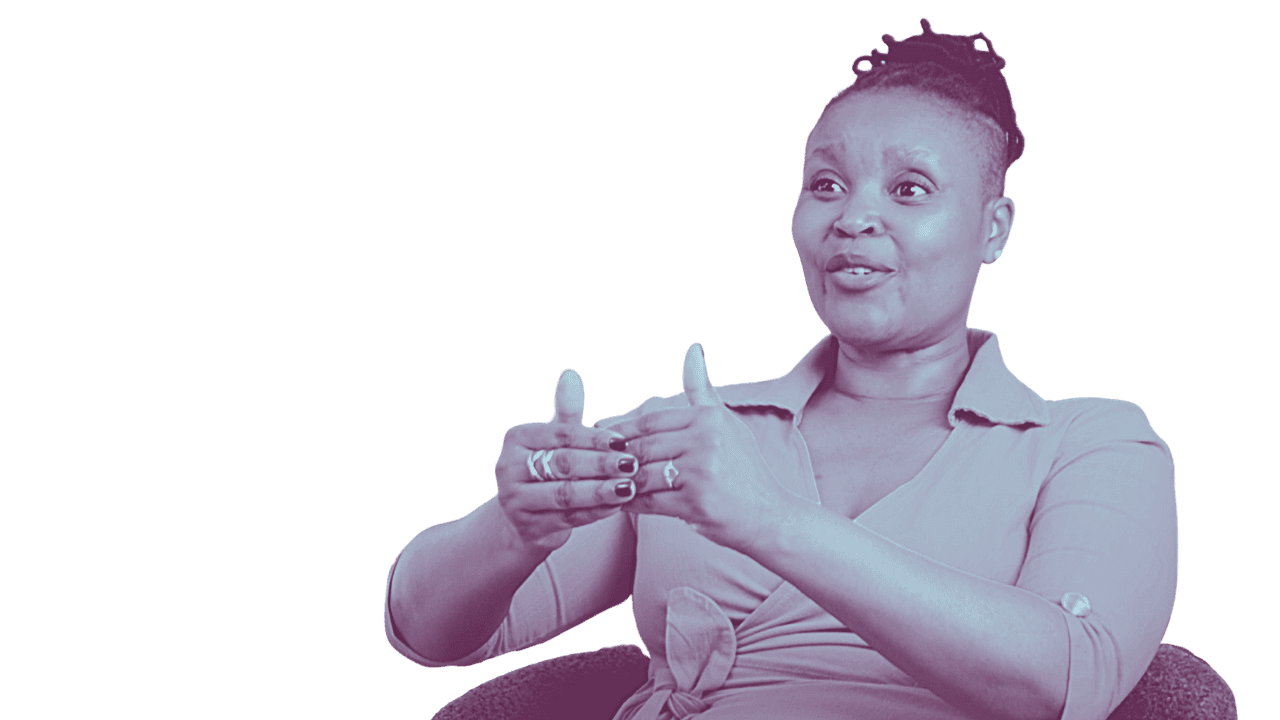 A woman with short hair styled in twists, wearing a light-colored button-up shirt, is sitting and speaking with expressive hand gestures.