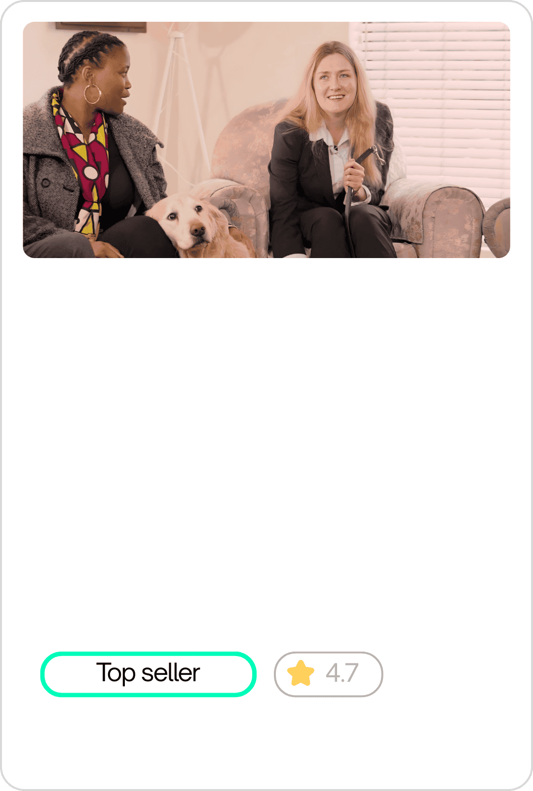 Two women sitting on a floral sofa in a room with white blinds. One woman, with light skin and long blond hair, is holding a leash and appears to be nervous or uncomfortable. The other woman, with dark skin and braids, is sitting beside a light-colored dog with a brown eye patch, looking at the woman with blond hair and smiling.