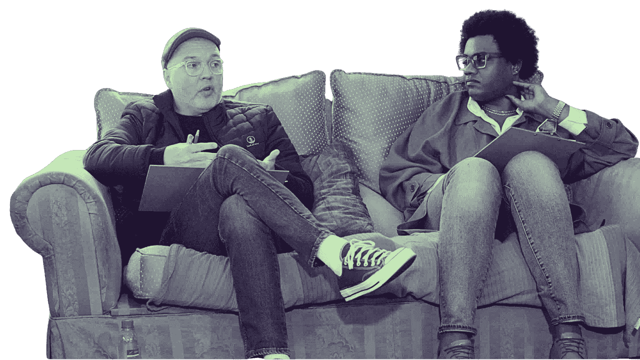Two men sitting on a sofa engaged in a conversation, one wearing glasses, a cap, and casual clothing, the other wearing glasses, an afro hairstyle, and casual fashion, with a black background.