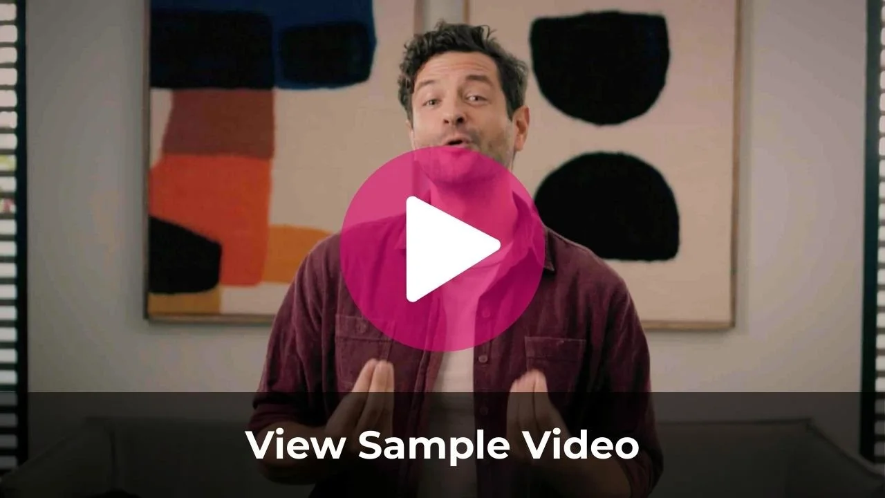 A man with dark curly hair and a beard, wearing a maroon jacket, stands in front of an abstract wall art with black, orange, red, and beige geometric shapes. He appears to be talking, with his hands raised and mouth open.