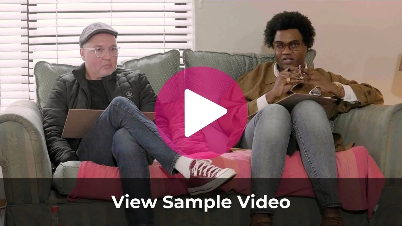 Two men sitting on a couch, one with glasses and a cap working on a laptop, the other with glasses and curly hair holding a phone, in a room with white blinds.