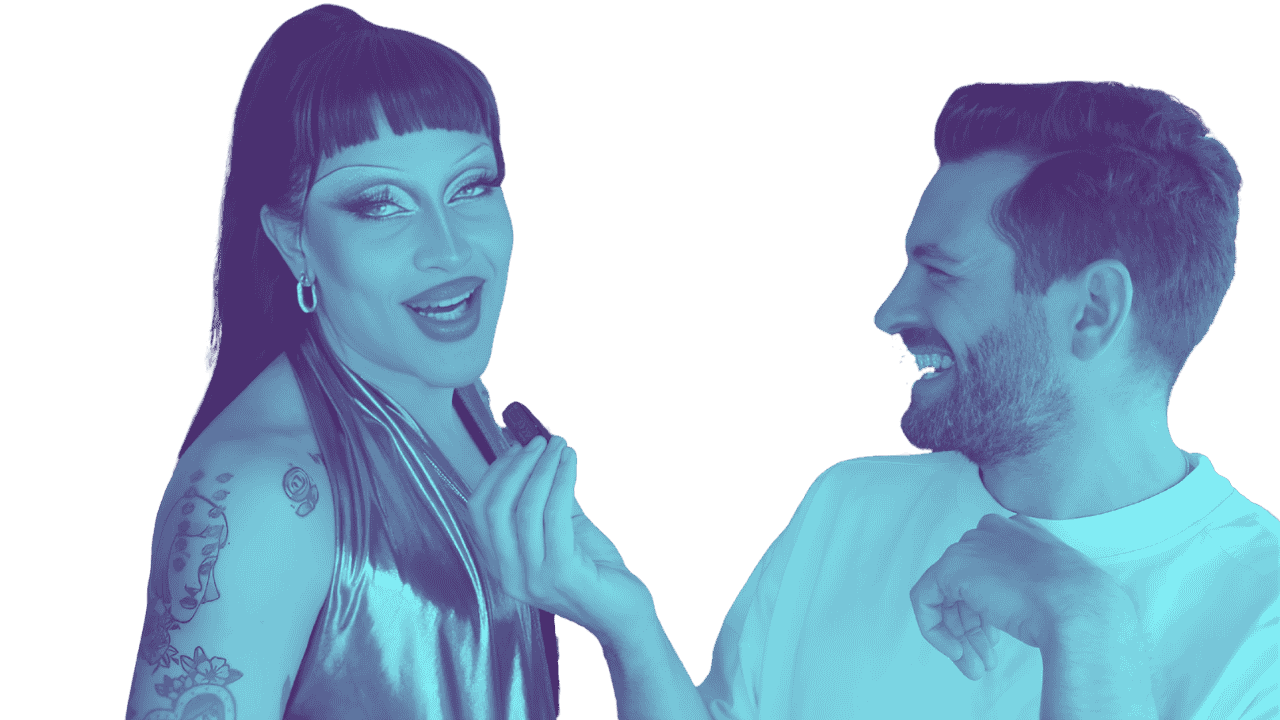 A drag queen with tattoos and dark hair talking to a man with a beard and light hair, both smiling and engaged in conversation.