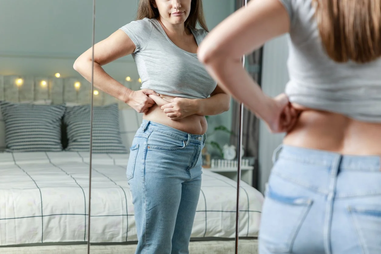 Displeased woman touching her belly looking in mirror at home