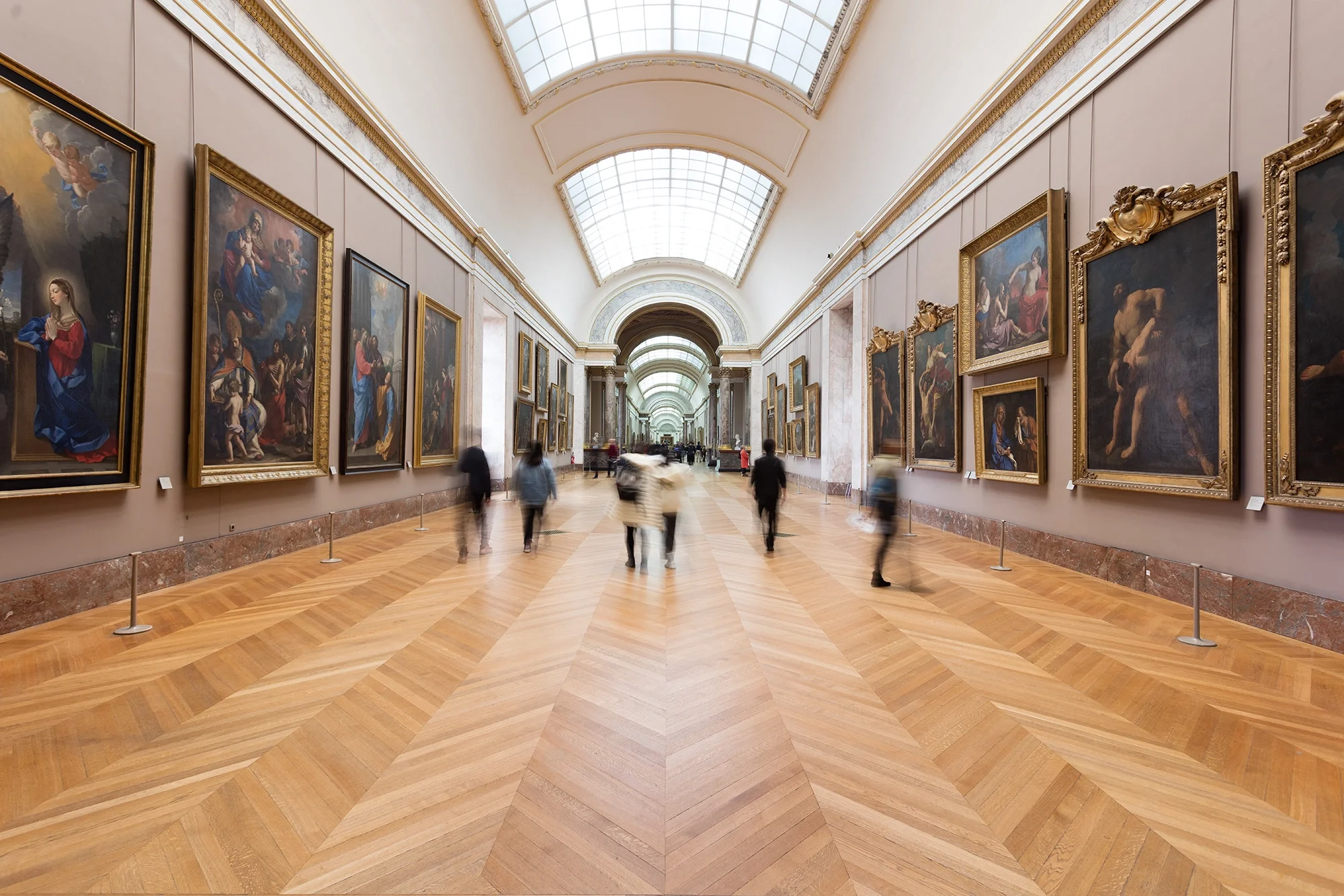 A spacious art gallery with tall ceilings and large arched windows allowing natural light. The walls are lined with framed classical paintings, and visitors are walking along the wooden floor, viewing the artwork.