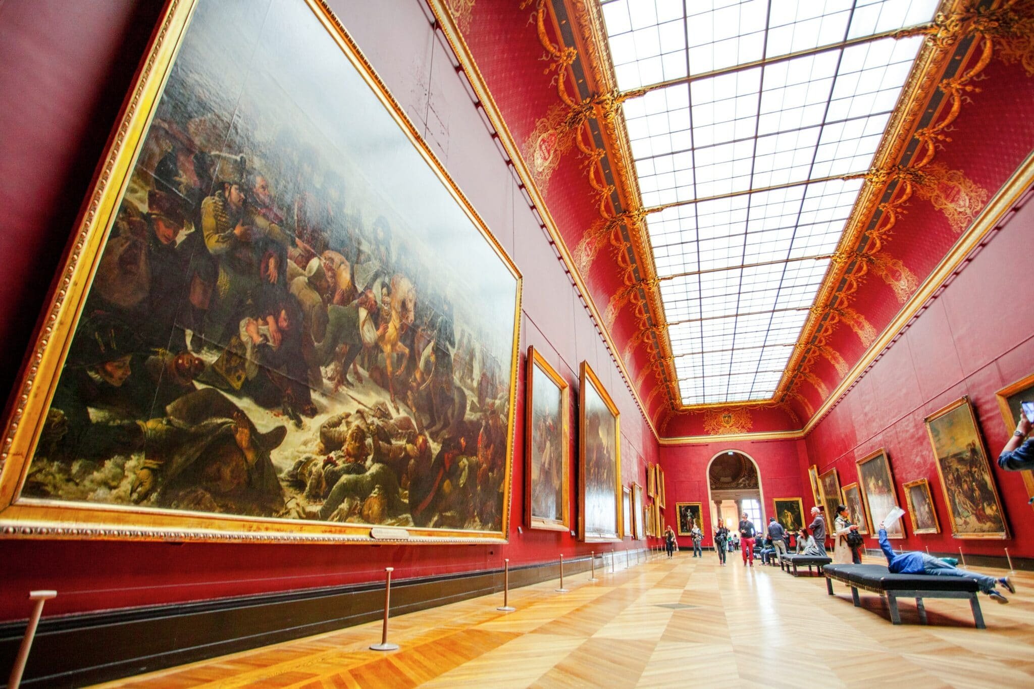 Interior of an art gallery with large, ornate paintings on red walls and a glass-roof ceiling. People are walking and sitting in the museum.