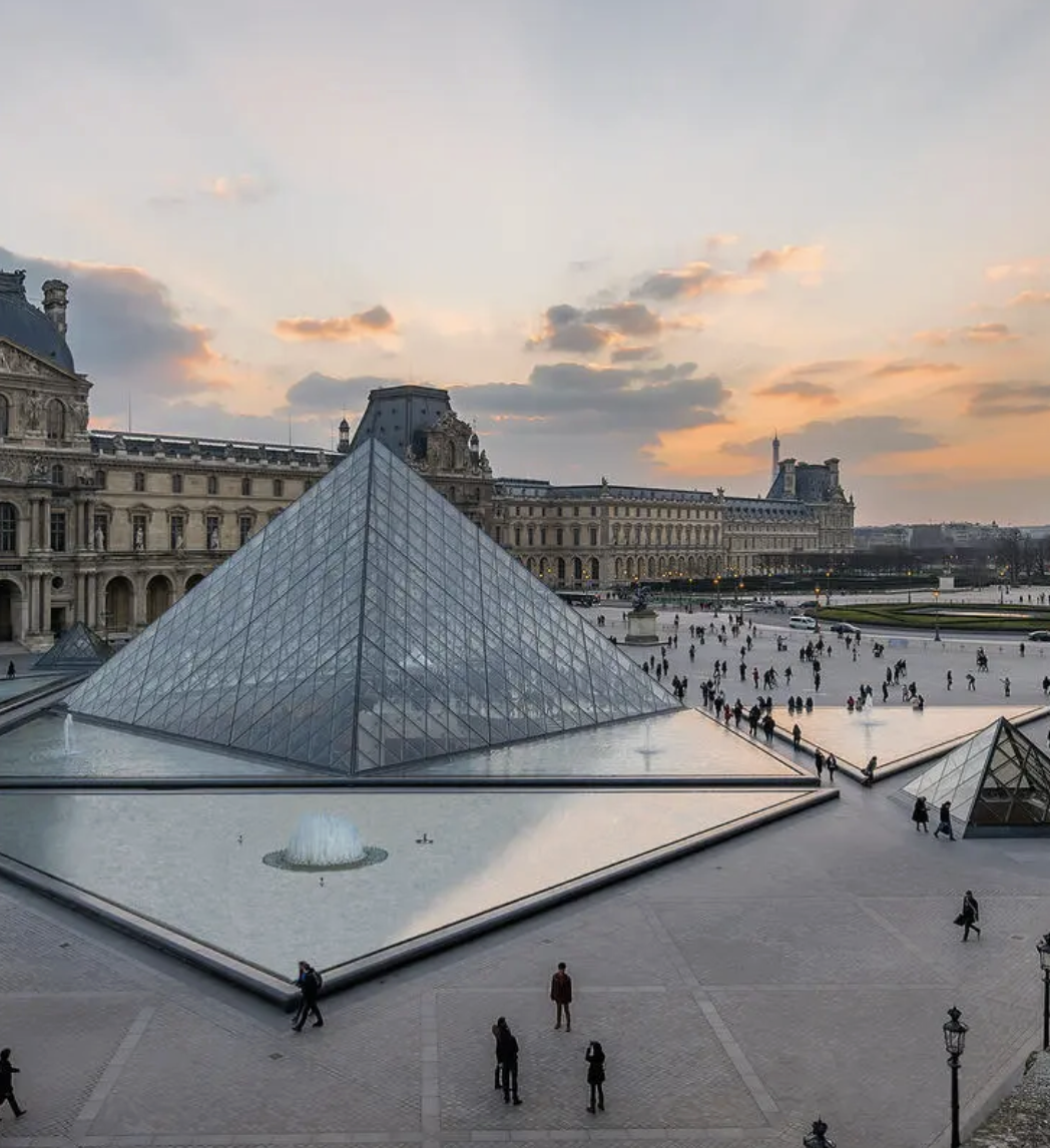 View of the Louvre Museum in Paris with Glass pyramids and a fountain in the foreground, the sky at sunset with scattered clouds and sunlight peeking through.