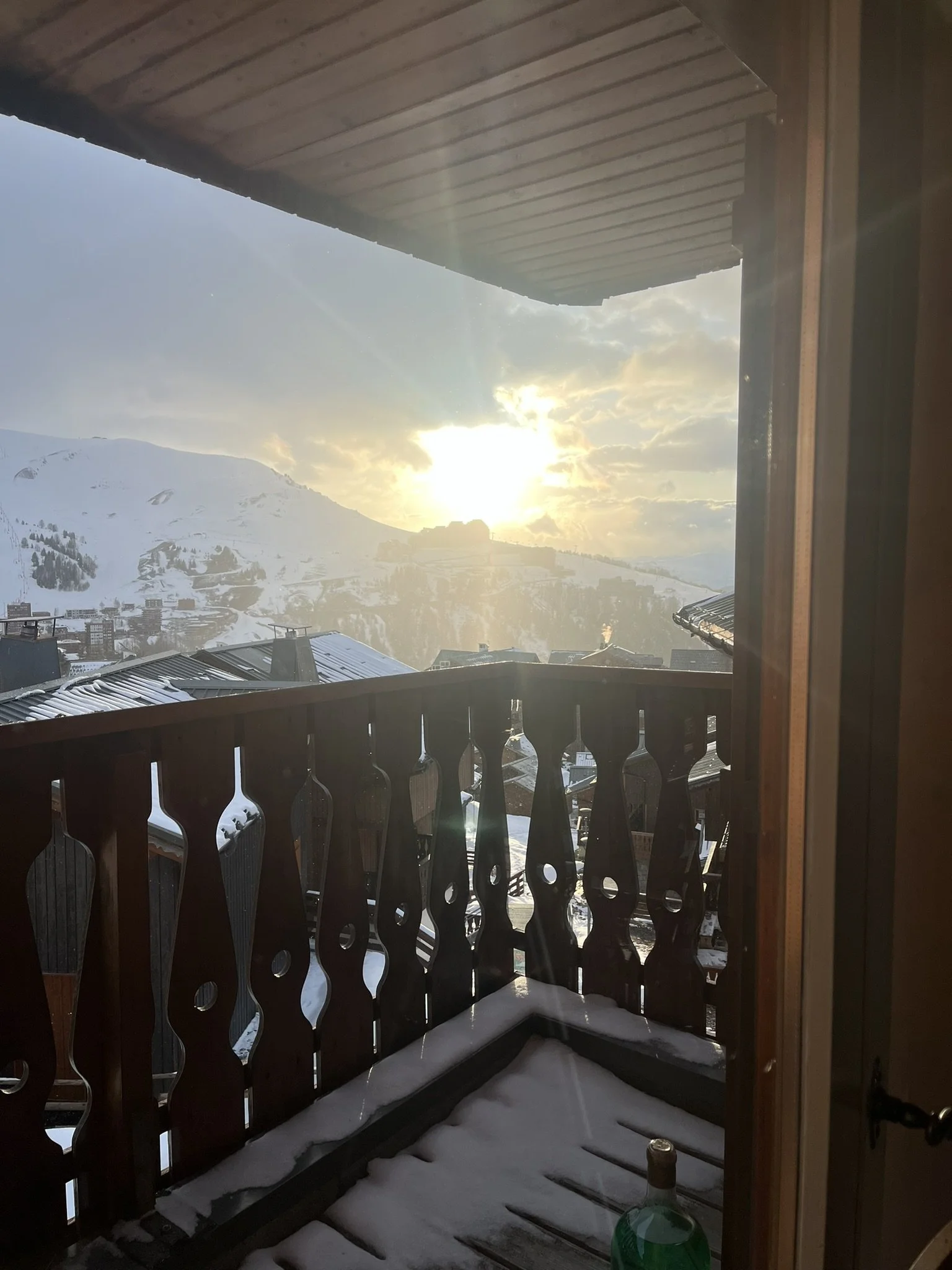 Vue d'un balcon en bois avec du neige, avec un paysage de montagnes enneigées et le soleil couchant derrière des nuages.