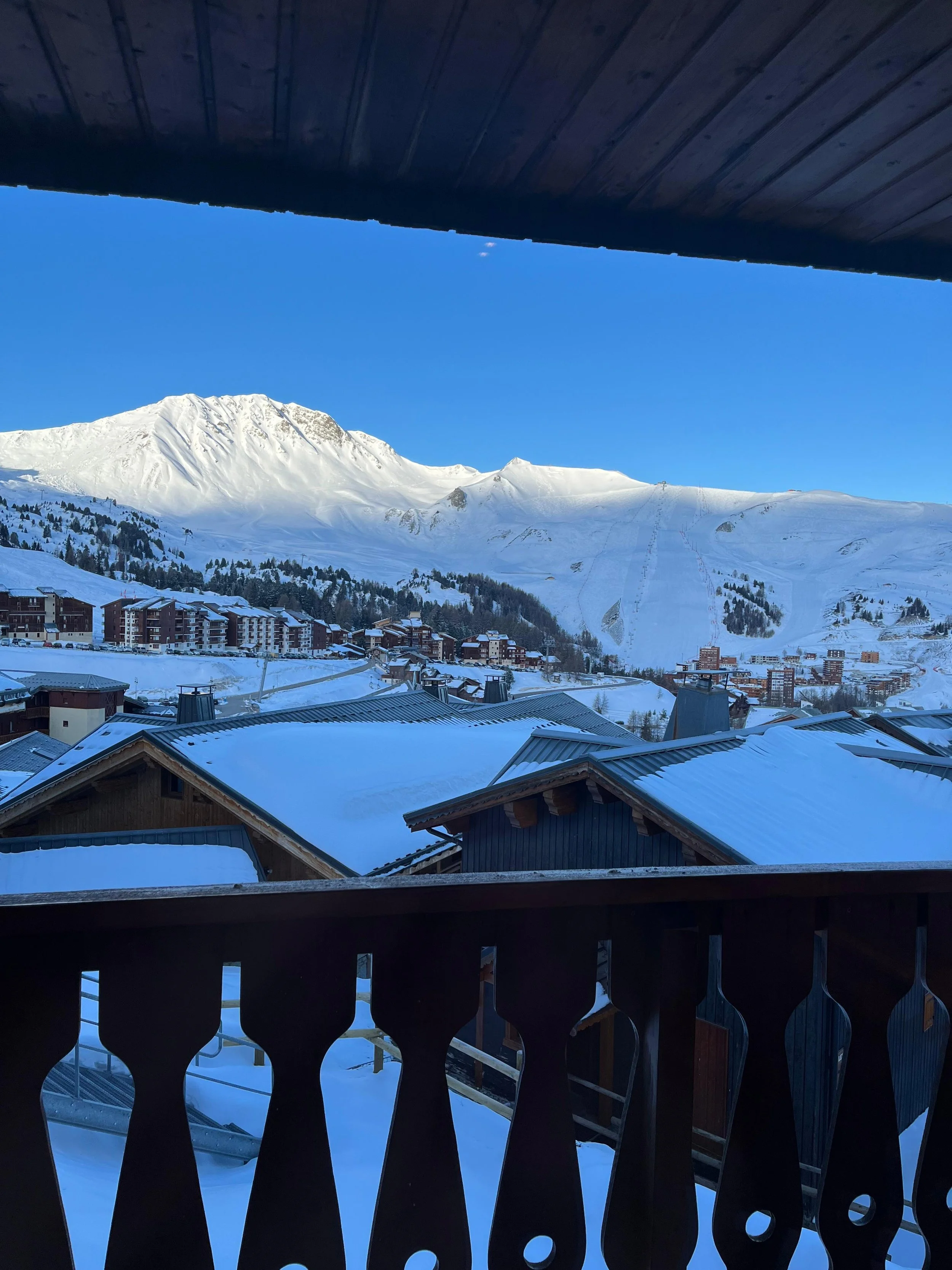 Vue sur une vallée enneigée avec des bâtiments en chalet, des montagnes enneigées au loin et un ciel clair, vue depuis un balcon en bois.