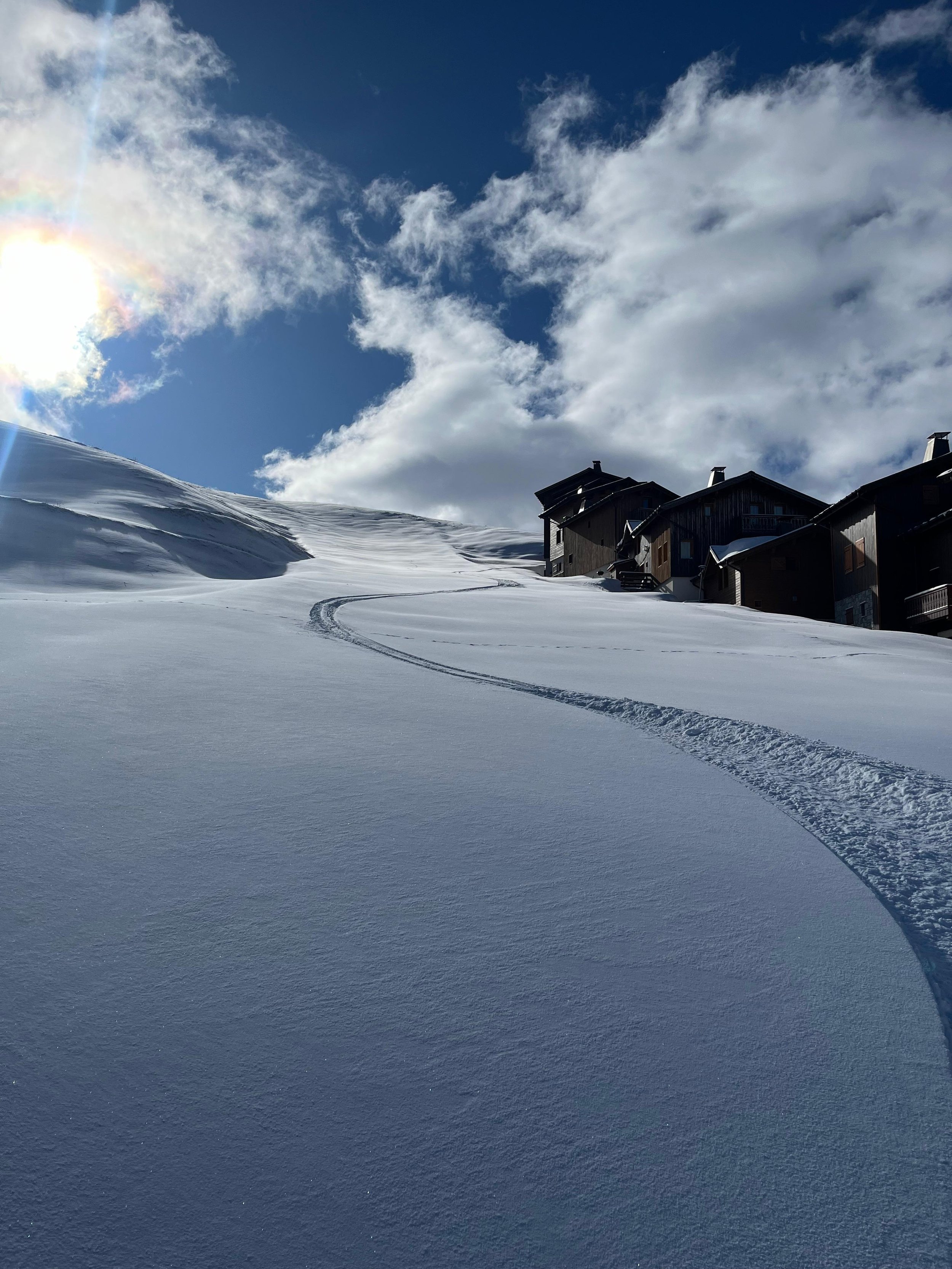 Paysage enneigé avec des maisons en bois, ciel nuageux, soleil brillant, traces de ski dans la neige.