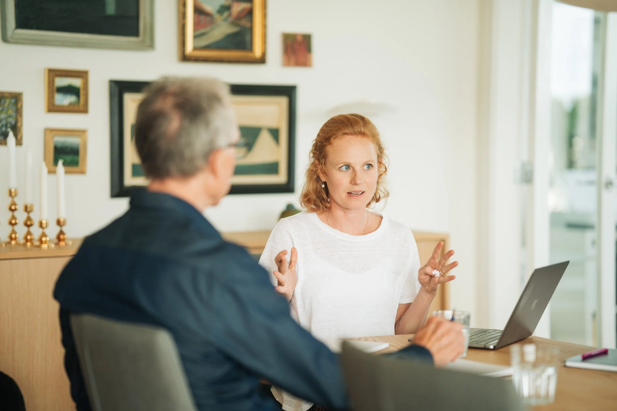 A woman with red hair and a white shirt gesturing while talking to a man with gray hair and glasses, sitting at a table with a laptop in a room decorated with framed artwork and candlesticks.