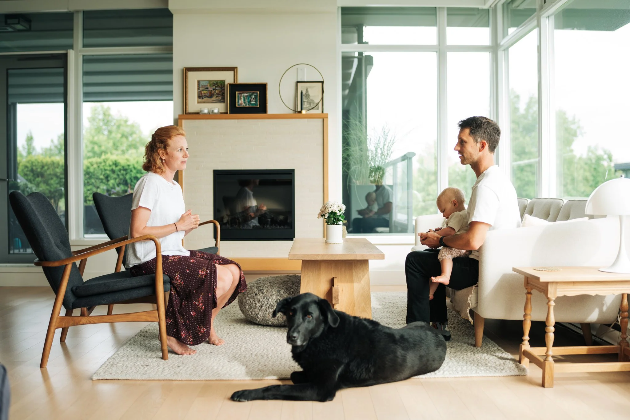 A woman and a man with a young child sitting on a white sectional sofa in a bright living room with large glass windows. The woman is sitting in a black armchair, facing the man, who is holding the child on his lap. A black dog is lying on a beige rug in the foreground. The room has a modern decor with a fireplace, framed pictures, and a large window overlooking greenery.