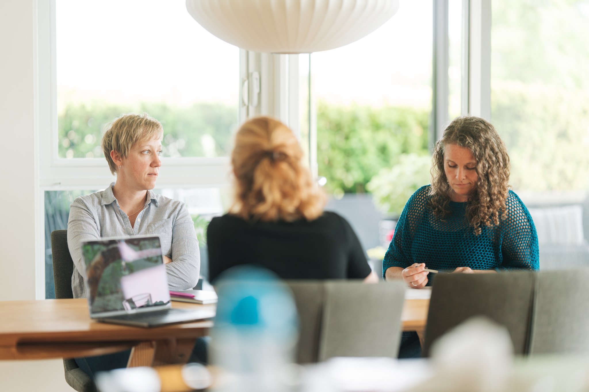 Three women sitting around a conference table in a bright room with large windows, engaged in a discussion or meeting.