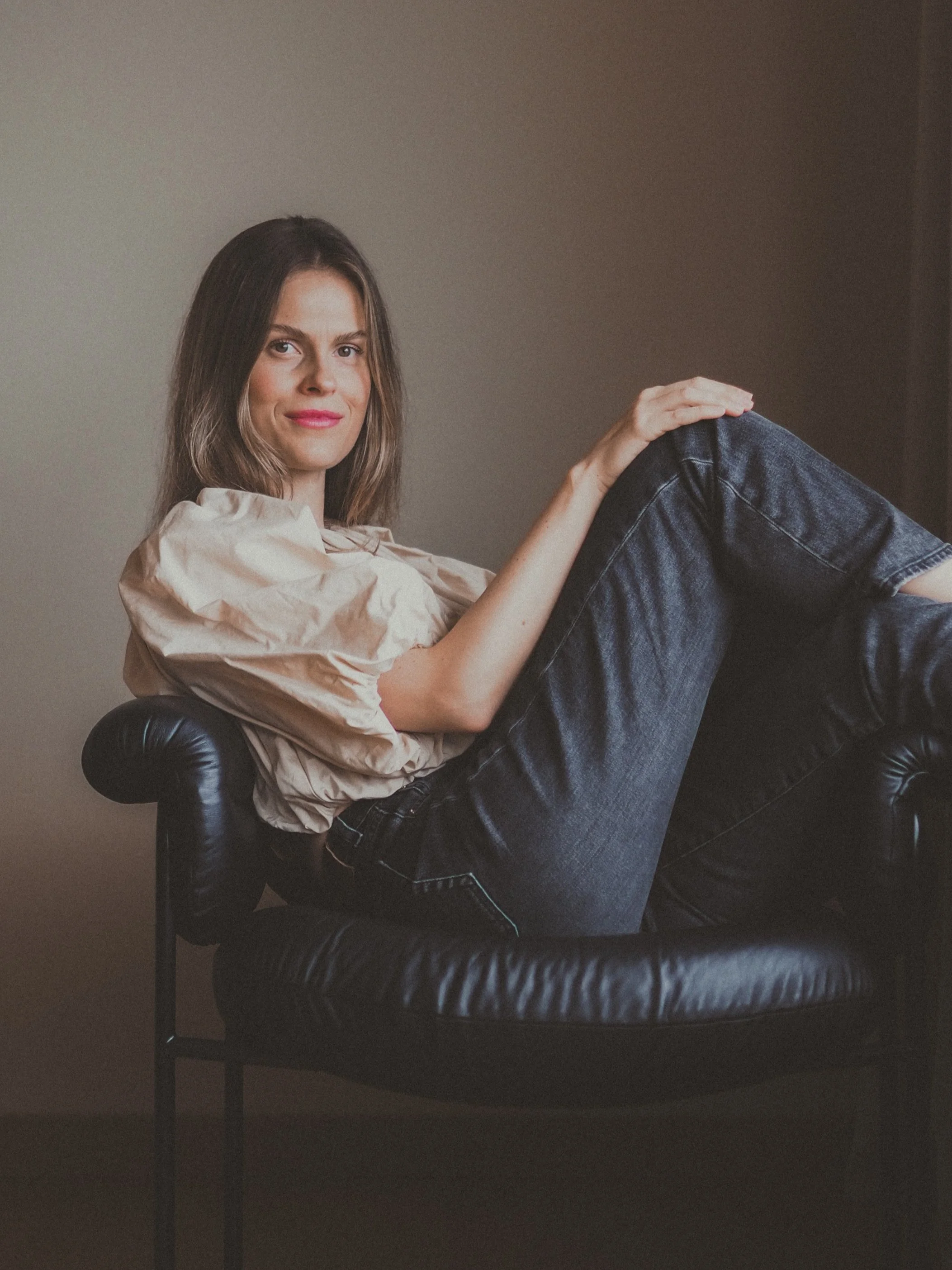 A young woman with shoulder-length brown hair and light makeup is sitting on a black leather armchair, smiling at the camera. She is wearing a beige blouse and dark jeans, with one leg propped up on the chair.