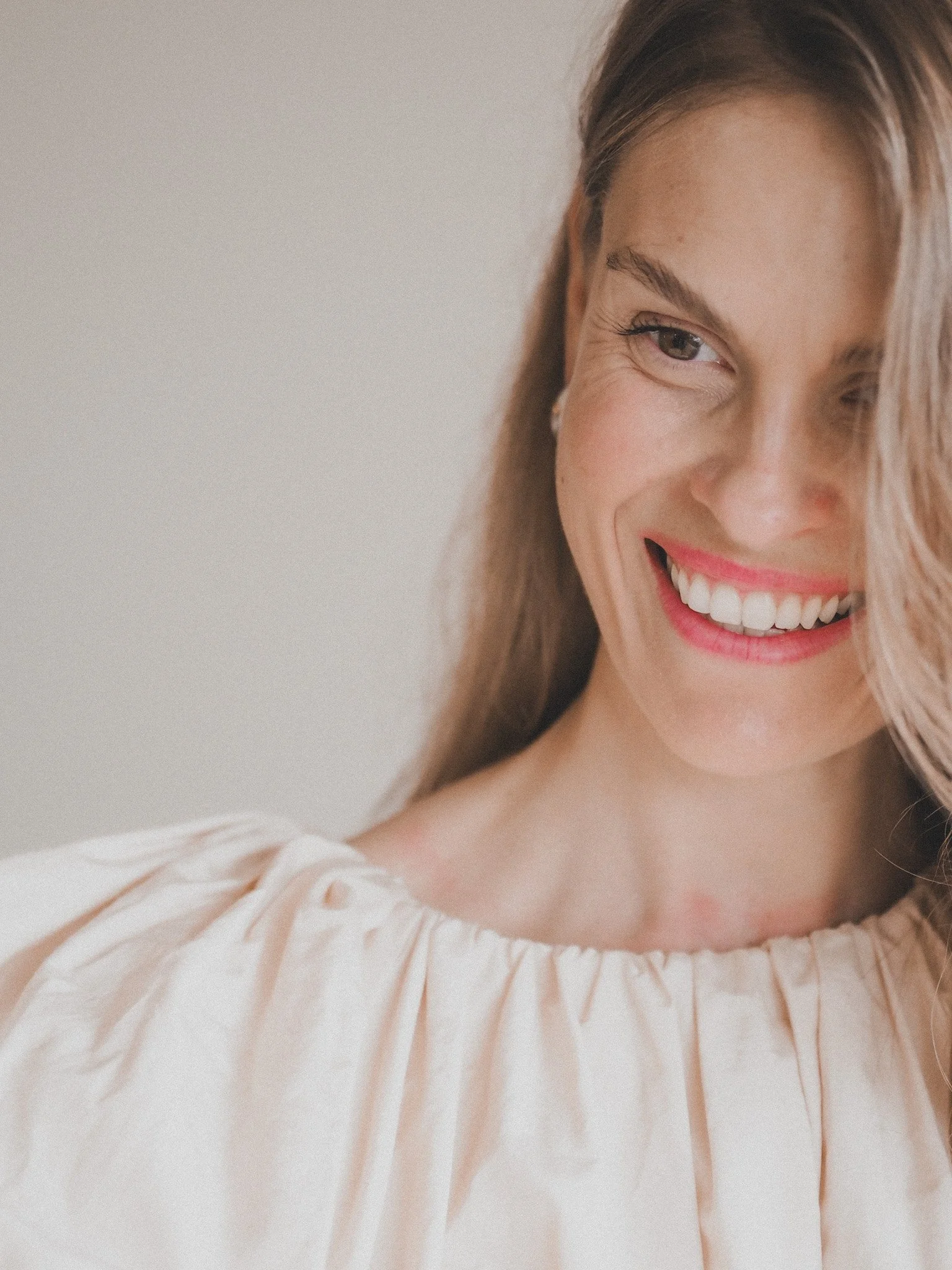 A woman with long, wavy hair smiling cheerfully, wearing a light-colored, gathered blouse.