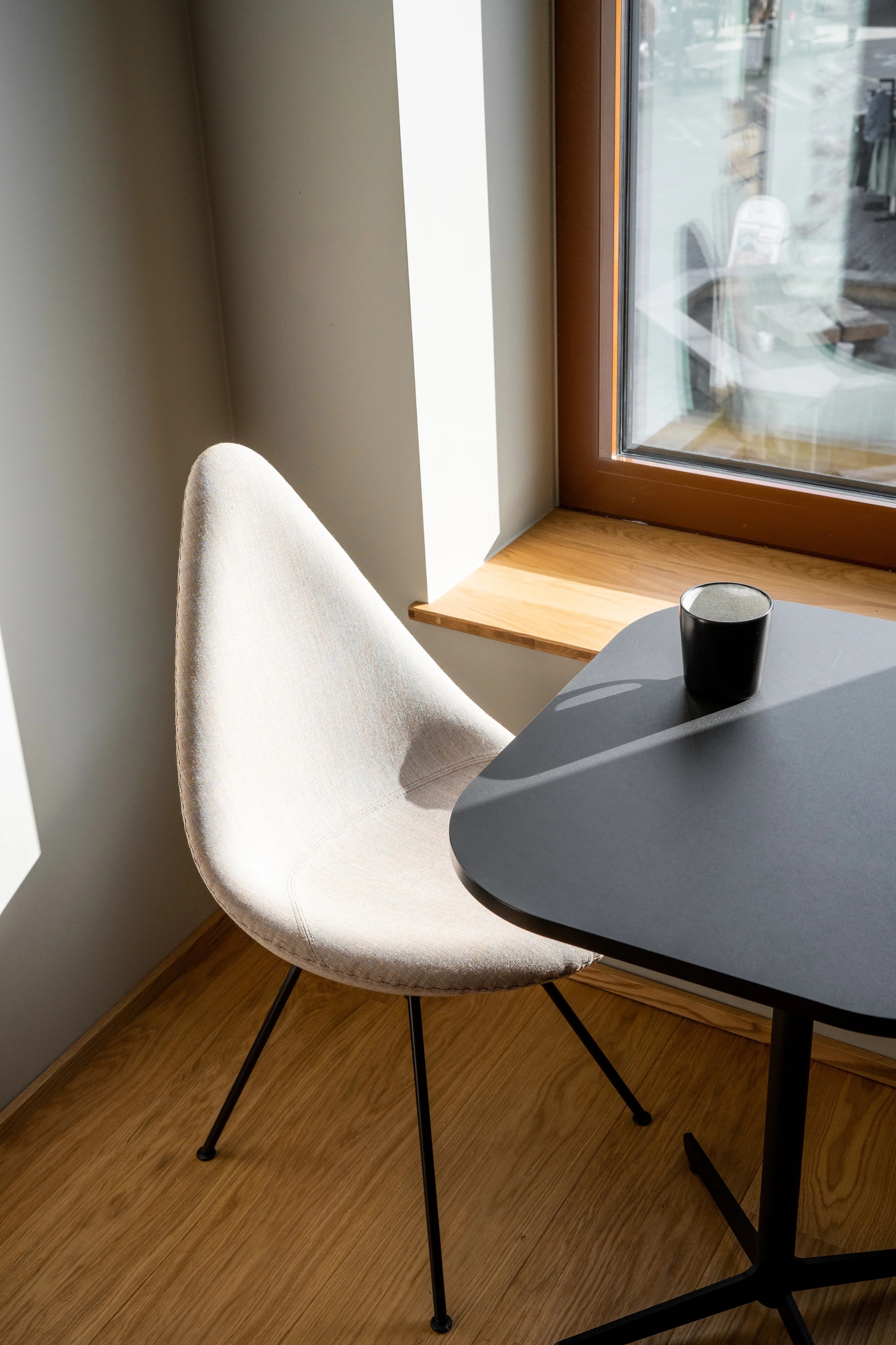 A white upholstered chair with black metal legs next to a dark gray table with a black cup on it, near a window letting in natural sunlight.