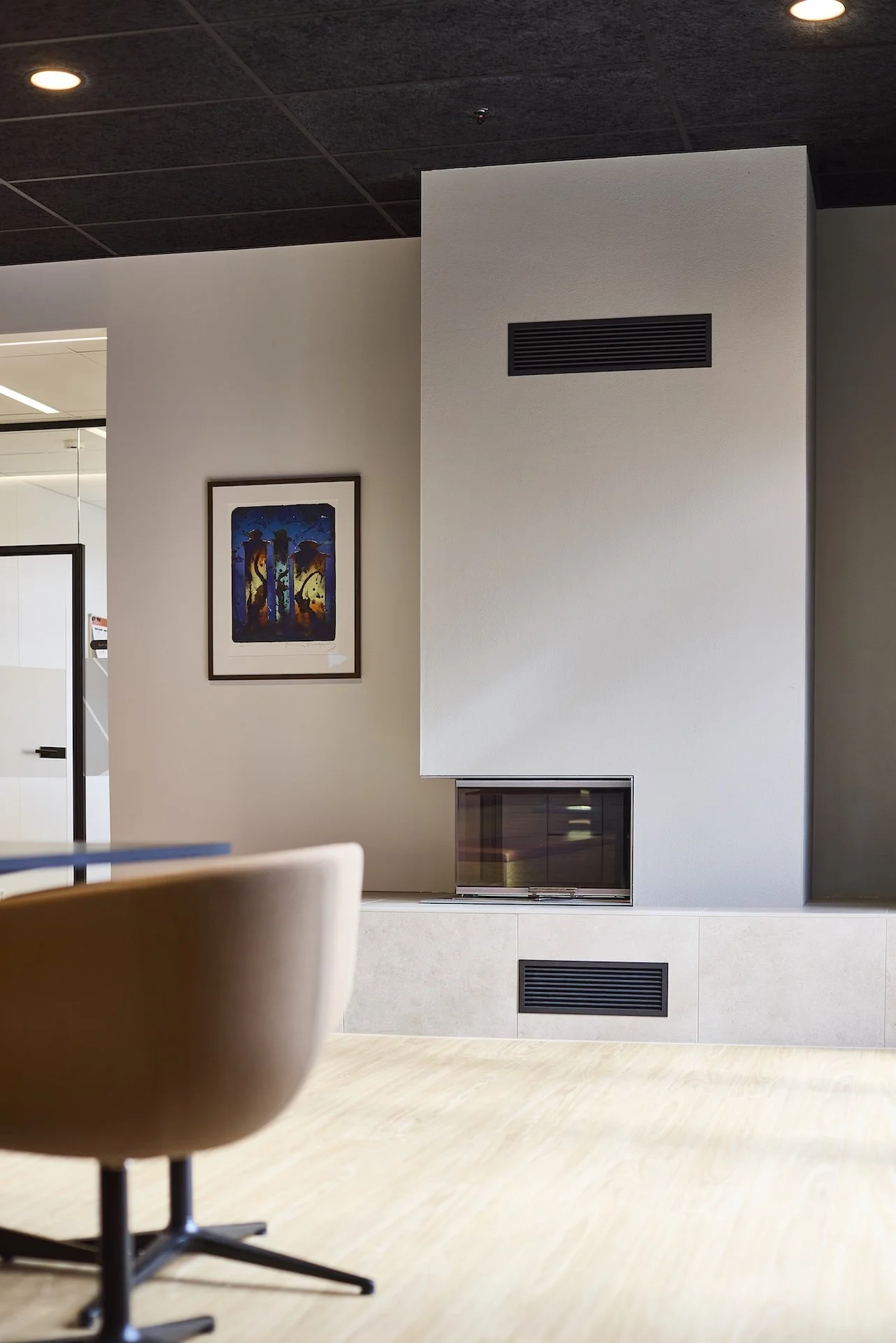 Modern office lounge with a beige chair, framed colorful abstract art on a white wall, and a sleek fireplace with vents.