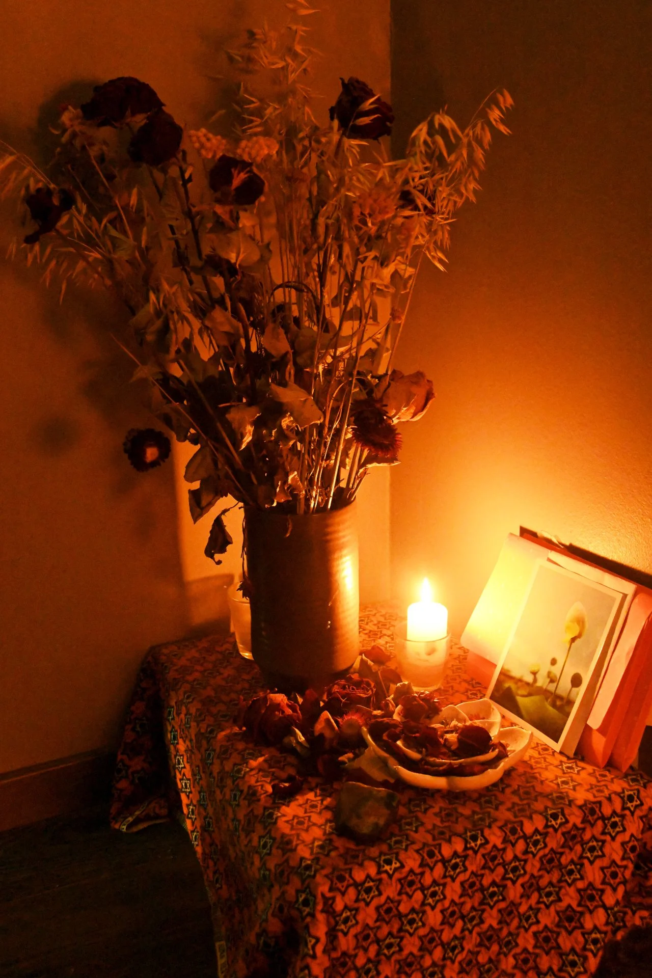 Vase contenant des fleurs séchées, bougie allumée, et photographies ou cartes posées sur une table recouverte d'un tissu avec motif géométrique, dans une pièce à l'éclairage tamisé.
