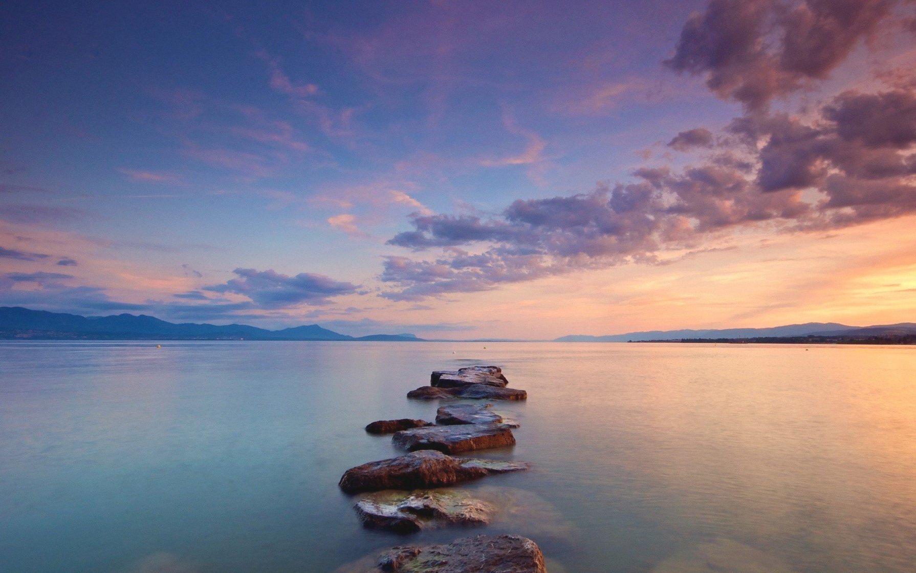 Scenic view of a lake with stepping stones leading into the water, surrounded by mountains and colorful sunset clouds.