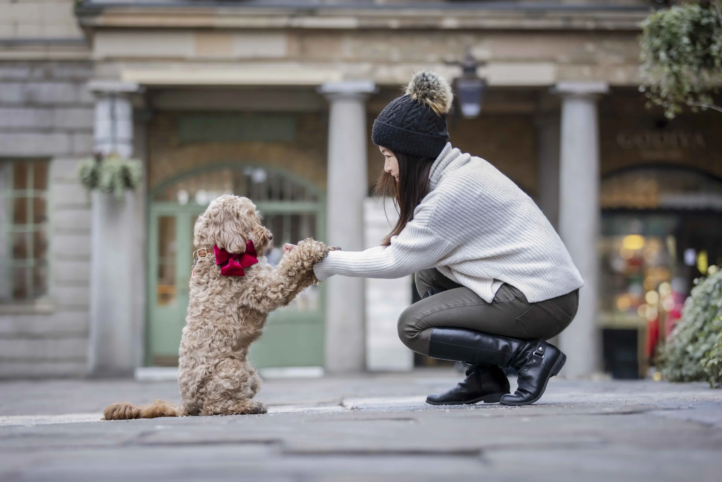 A woman in a gray sweater, black boots, and a black knit hat with a pom-pom, crouching down, holding hands with a small curly-haired dog wearing a red bow, on a city street with buildings and storefronts in the background.