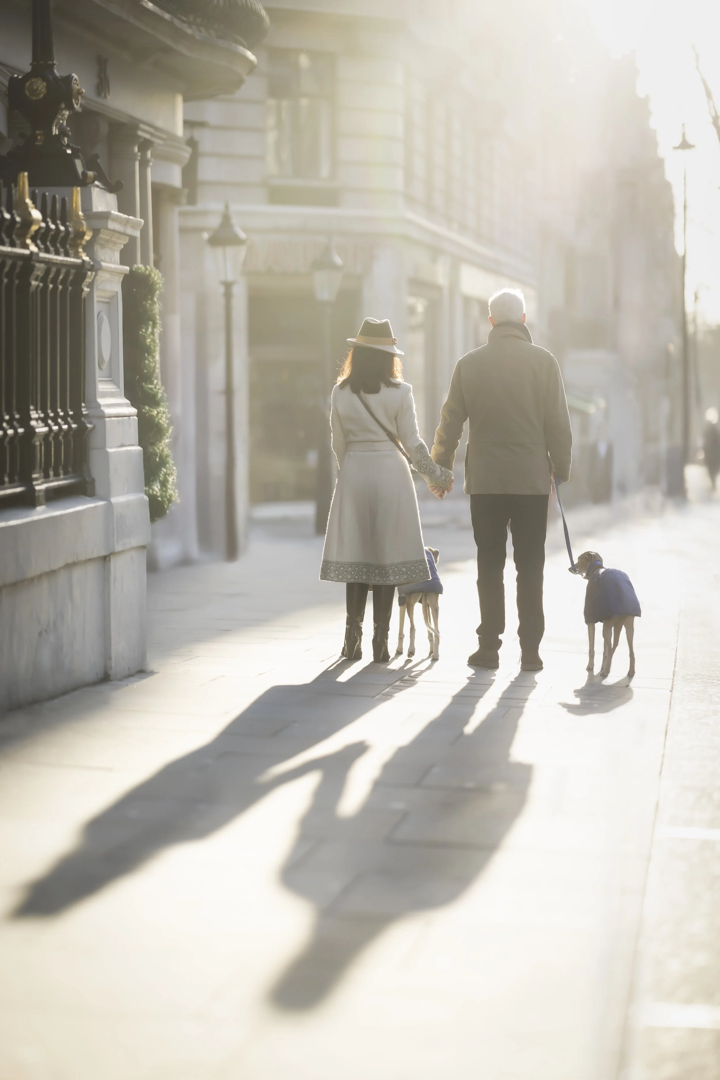 Couple holding hands while walking two Whippets through Mayfair, London, in the early morning light during a family and dog photoshoot.