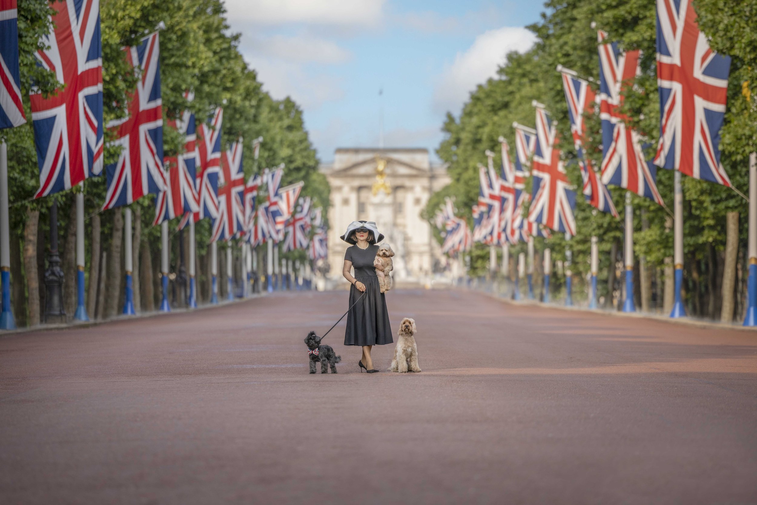 Woman in a wide-brimmed hat standing with three small rescue dogs on The Mall, surrounded by Union flags with Buckingham Palace in the background.