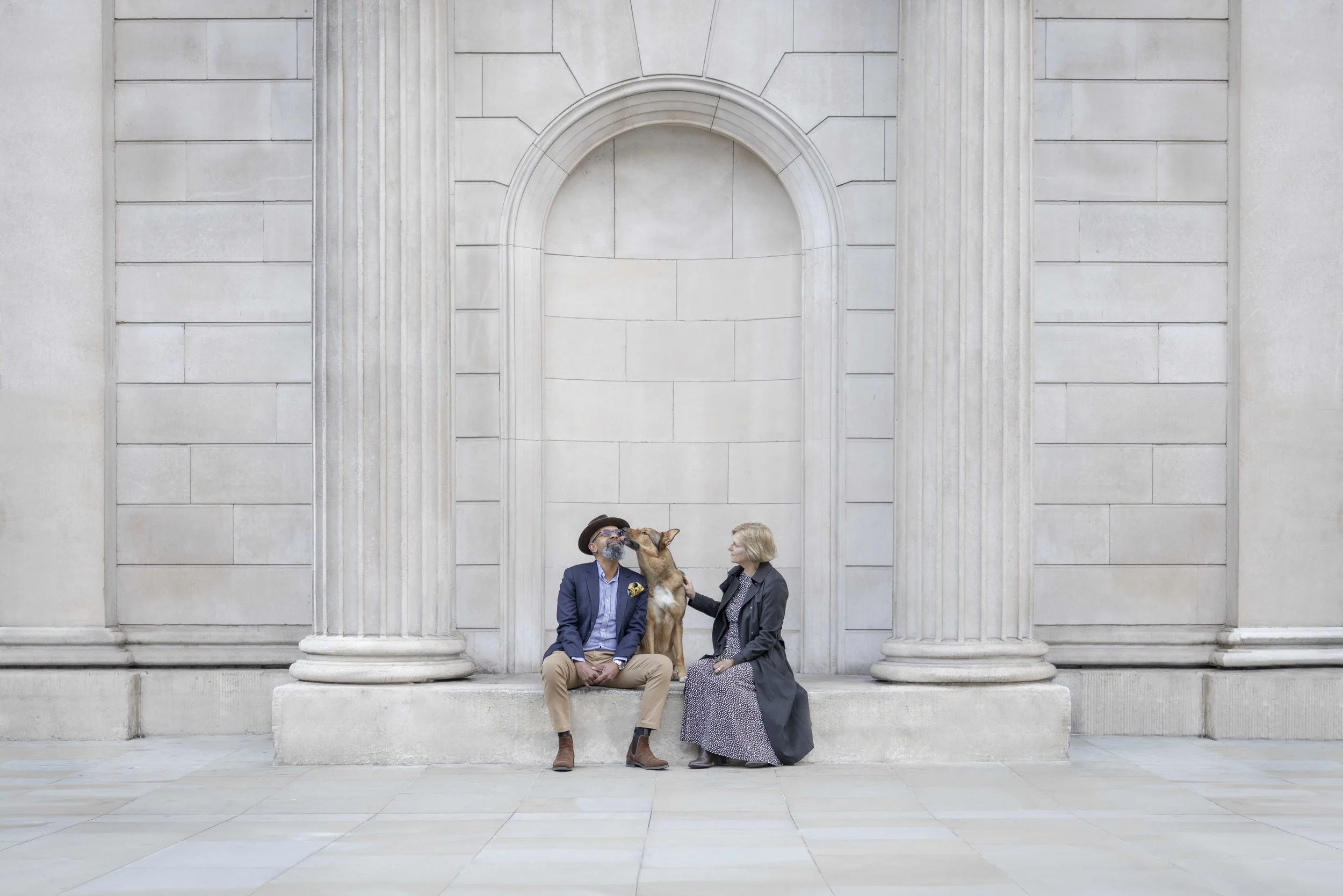 Couple sitting with their German Shepherd beneath grand stone columns in the City of London during a bespoke dog photography session.