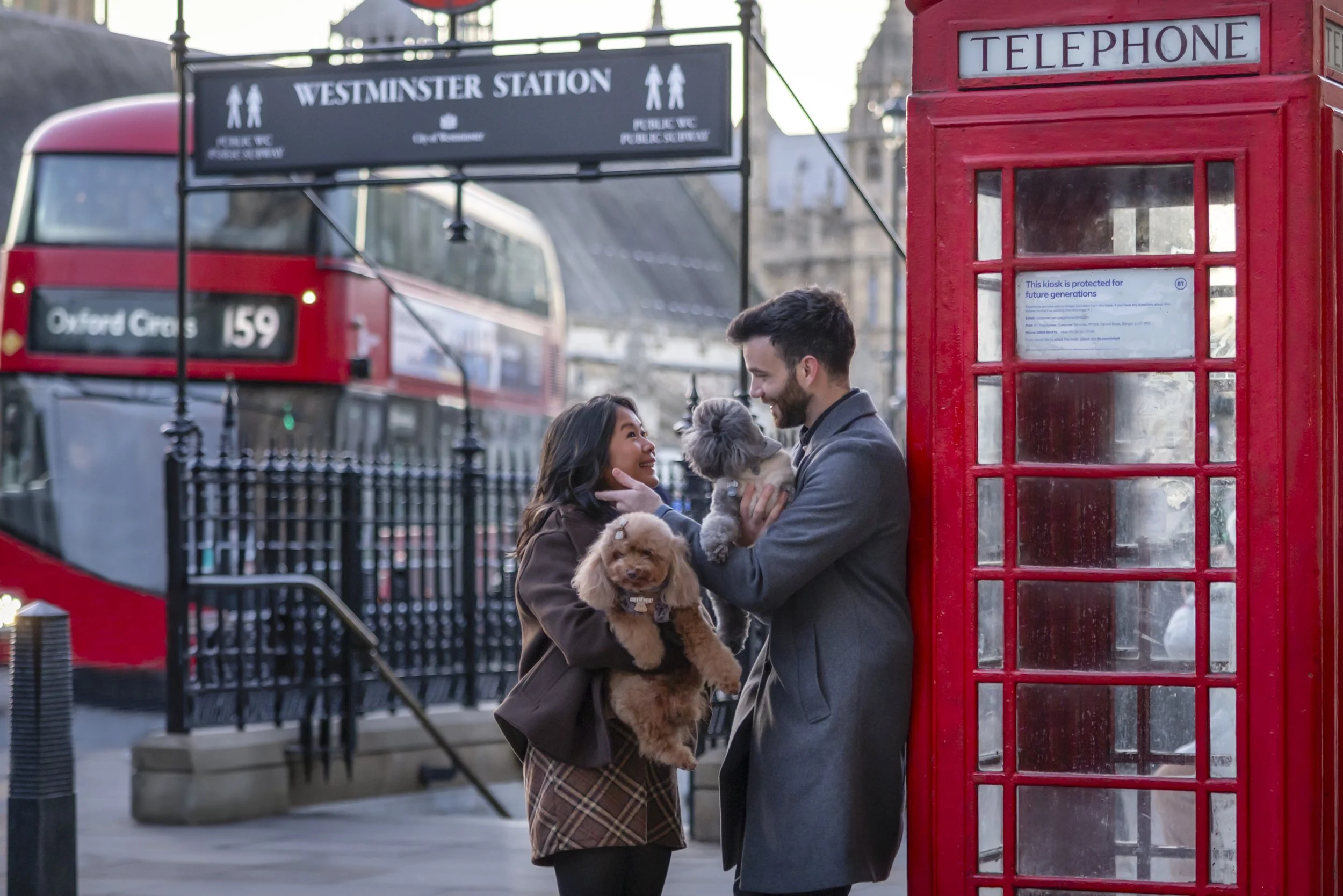 Couple holding their two Toy Poodles beside a red telephone box outside Westminster Station during a London dog photography session.