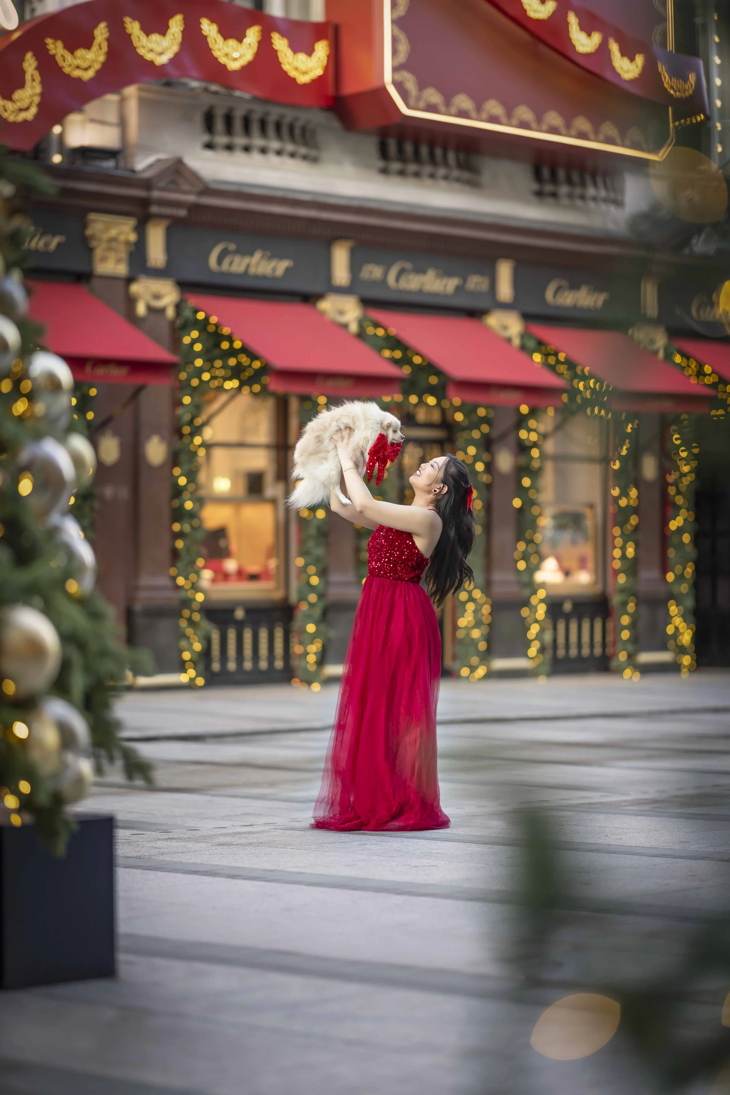 Woman in a red dress holding her small white Pomeranian beneath Christmas decorations outside Cartier on New Bond Street, Mayfair, London.