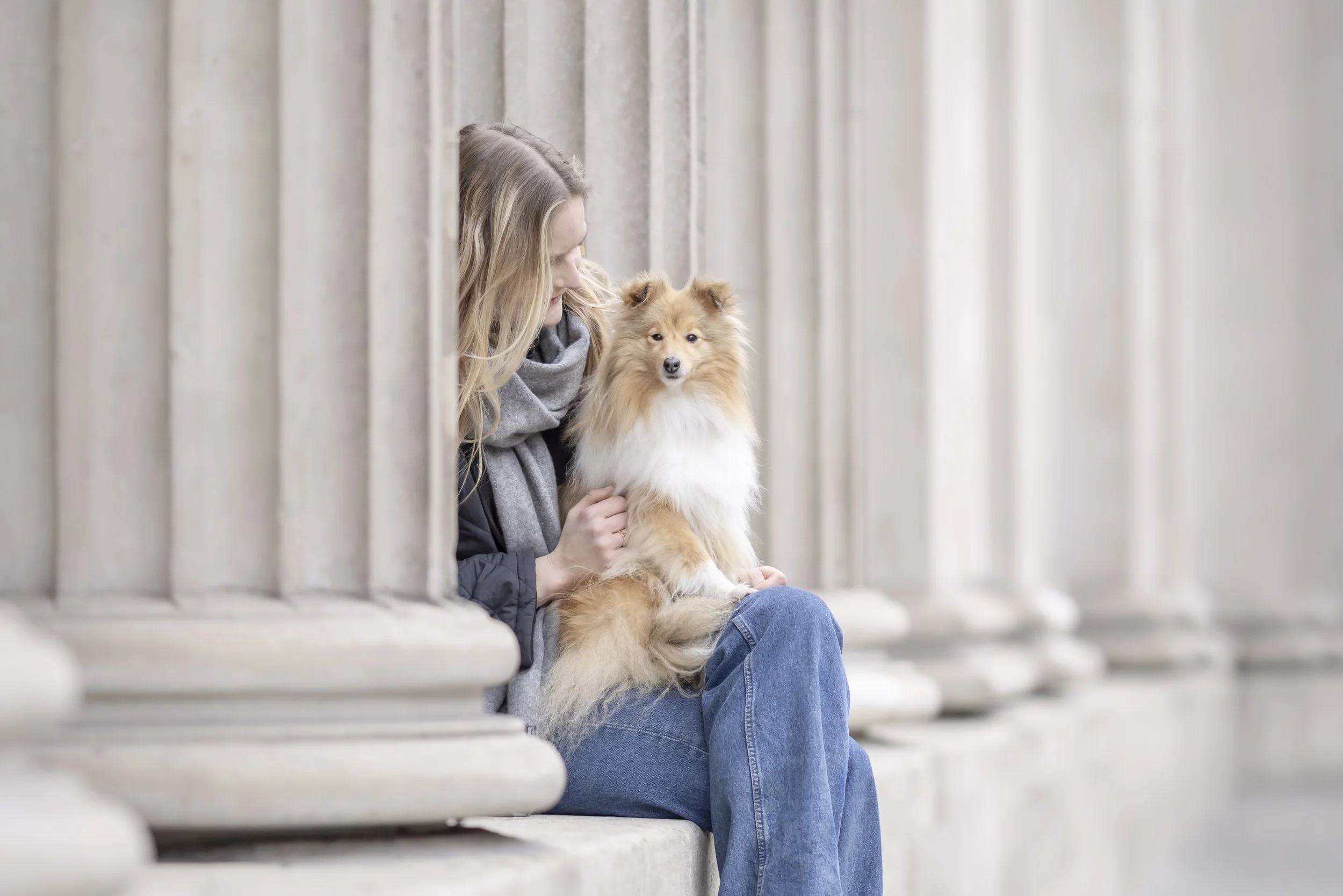 Woman seated beneath grand stone columns in the City of London holding her sable and white Shetland Sheepdog, Callie, during a natural pet and owner photography session.