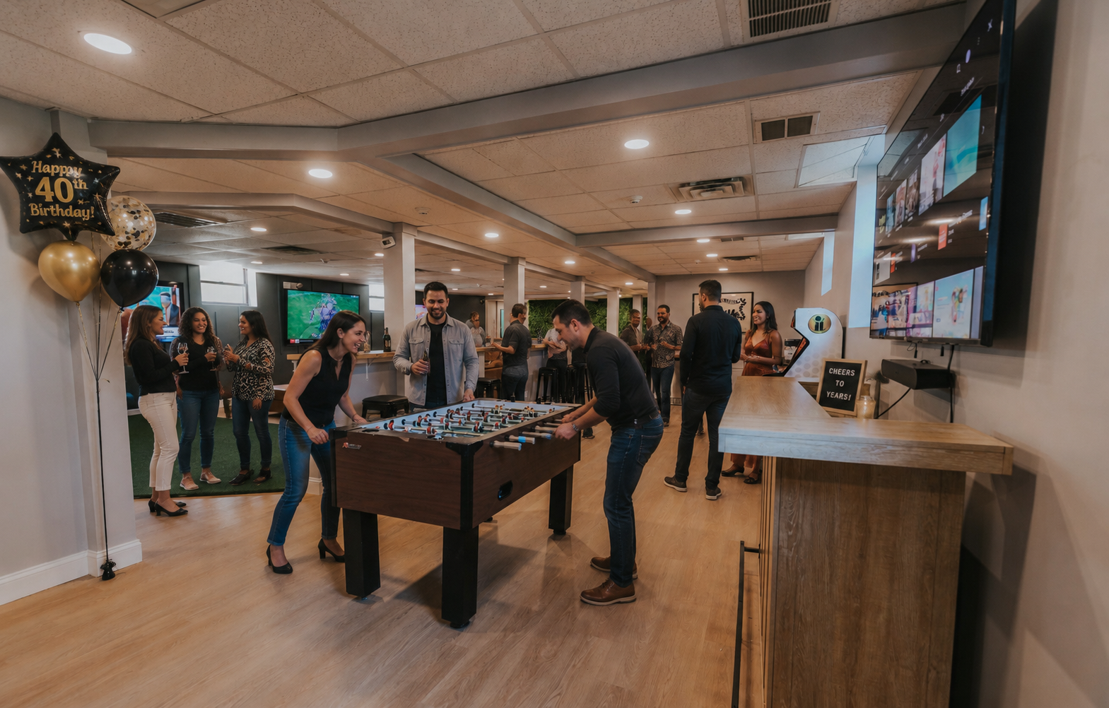 People celebrating a 40th birthday party in a bar or lounge, with some playing foosball, others socializing with drinks, and balloons and a sign indicating the celebration.