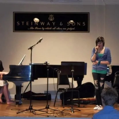 A new student performing live for the first time singing a beautiful arrangement. of one of her favorite songs while accompanied by a piano student on the keys. 