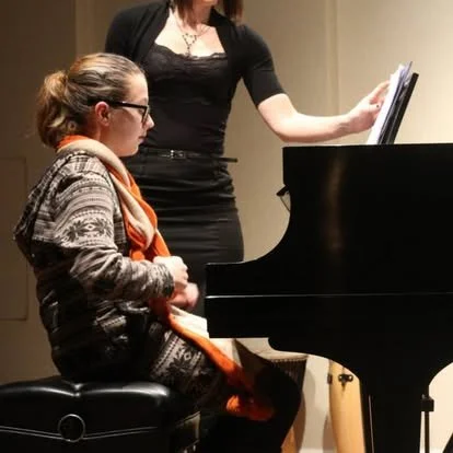A young girl with glasses and a ponytail sits at a piano, while a woman stands next to her, holding sheet music.