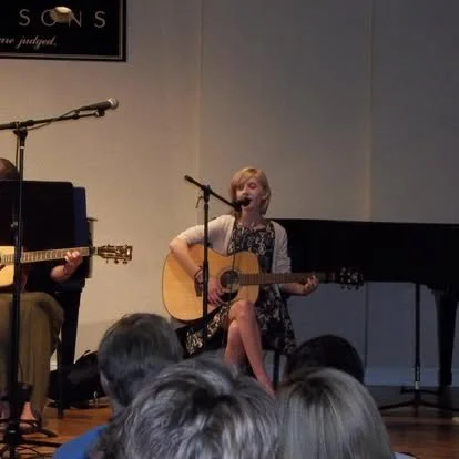 Student Anna singing and playing acoustic guitar while accompanied by Ms. Brown on vocals and acoustic guitar.