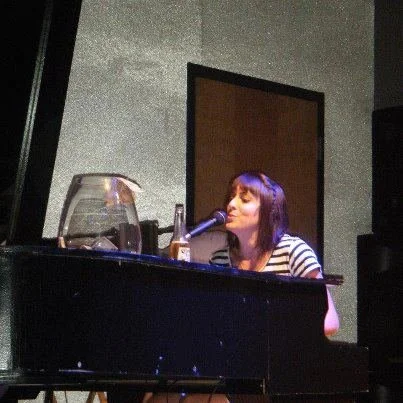 A woman with dark hair singing into a microphone at a black grand piano on a stage, with a glass of water and some bottles nearby.