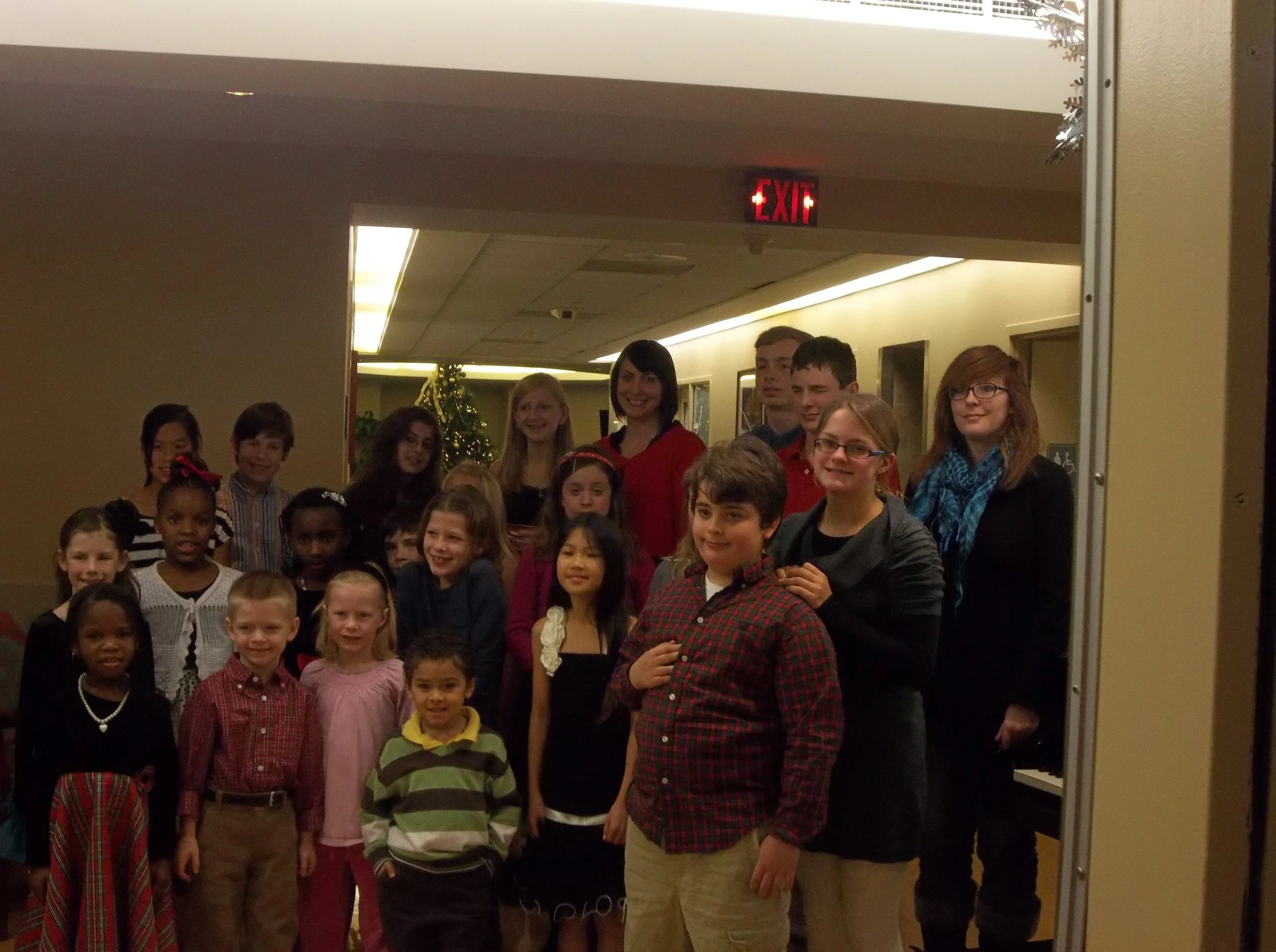 A group of children and adults gathered together for a photo inside a building, with a decorated Christmas tree in the background.