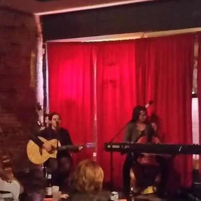 Two Prestige Performers instructors on stage at a benefit concert playing and singing a duet with cello and acoustic guitar