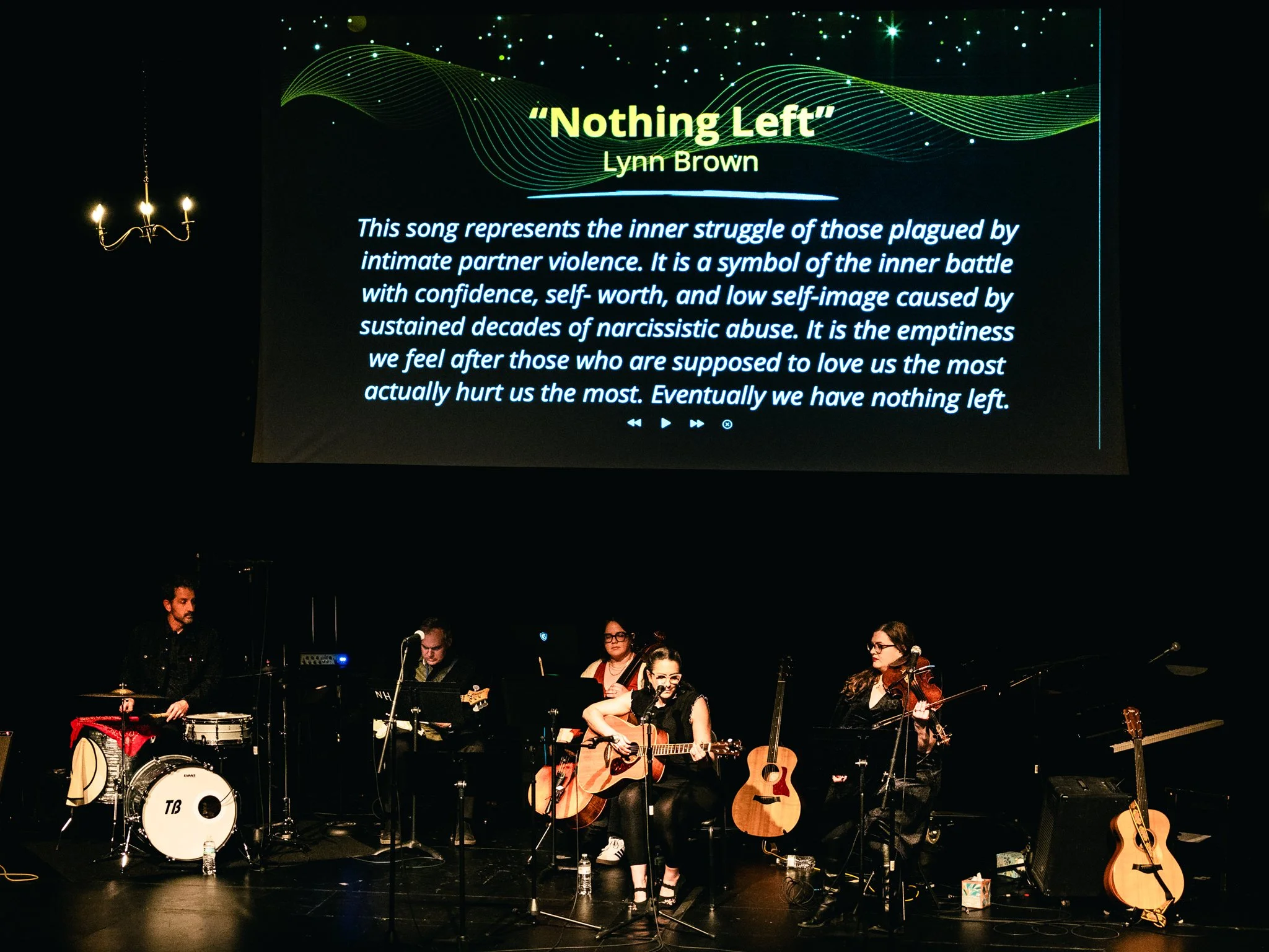 A musical performance on stage featuring a band with a woman singing and playing guitar, other musicians with instruments, and a large screen displaying the song title 'Nothing Left' by Lynn Brown along with a description of the song's meaning.