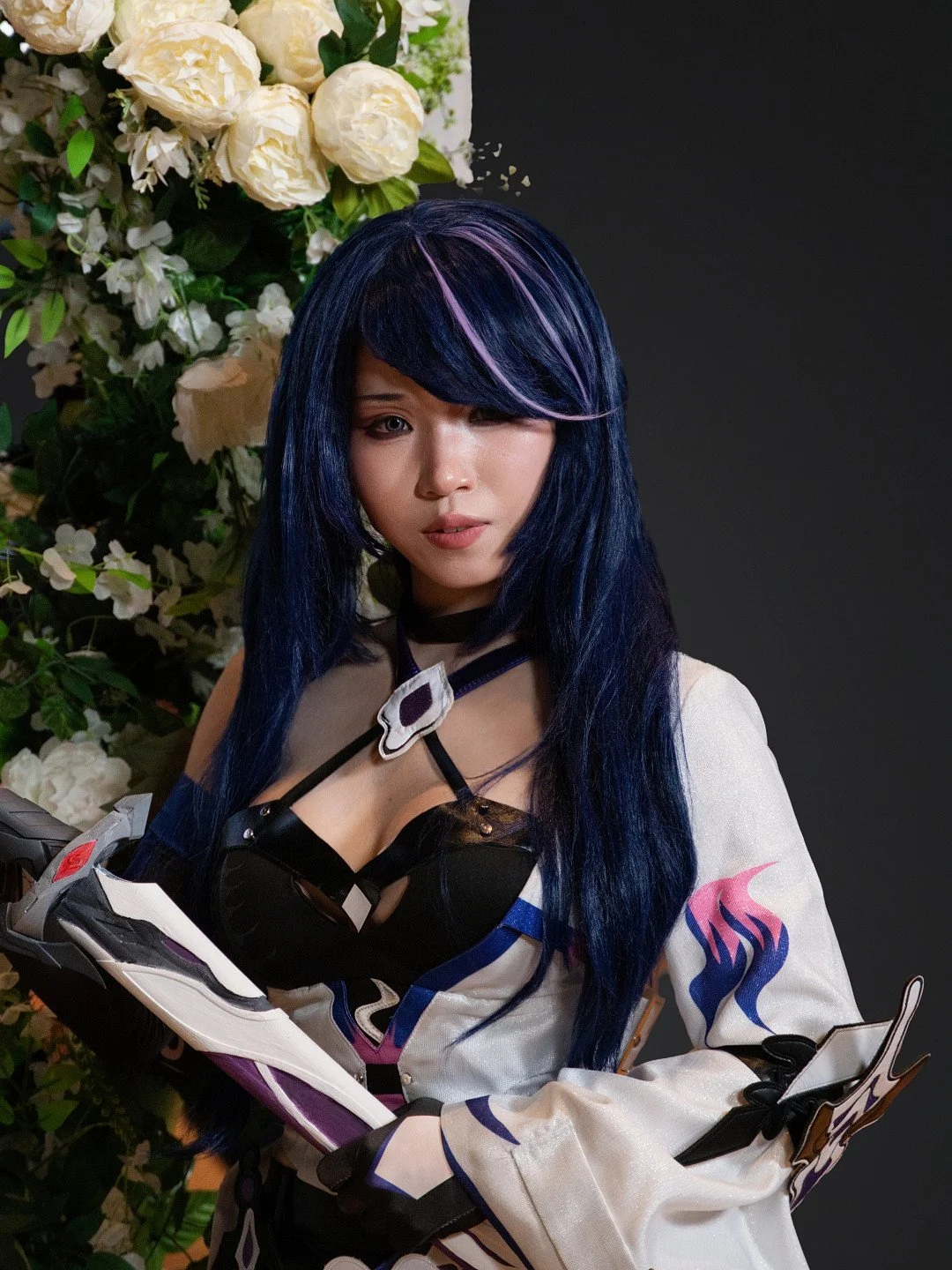 A woman with long blue hair and pink highlights, wearing a black and white outfit with gloves, holding a motorcycle helmet, standing next to a bouquet of white flowers against a dark background.